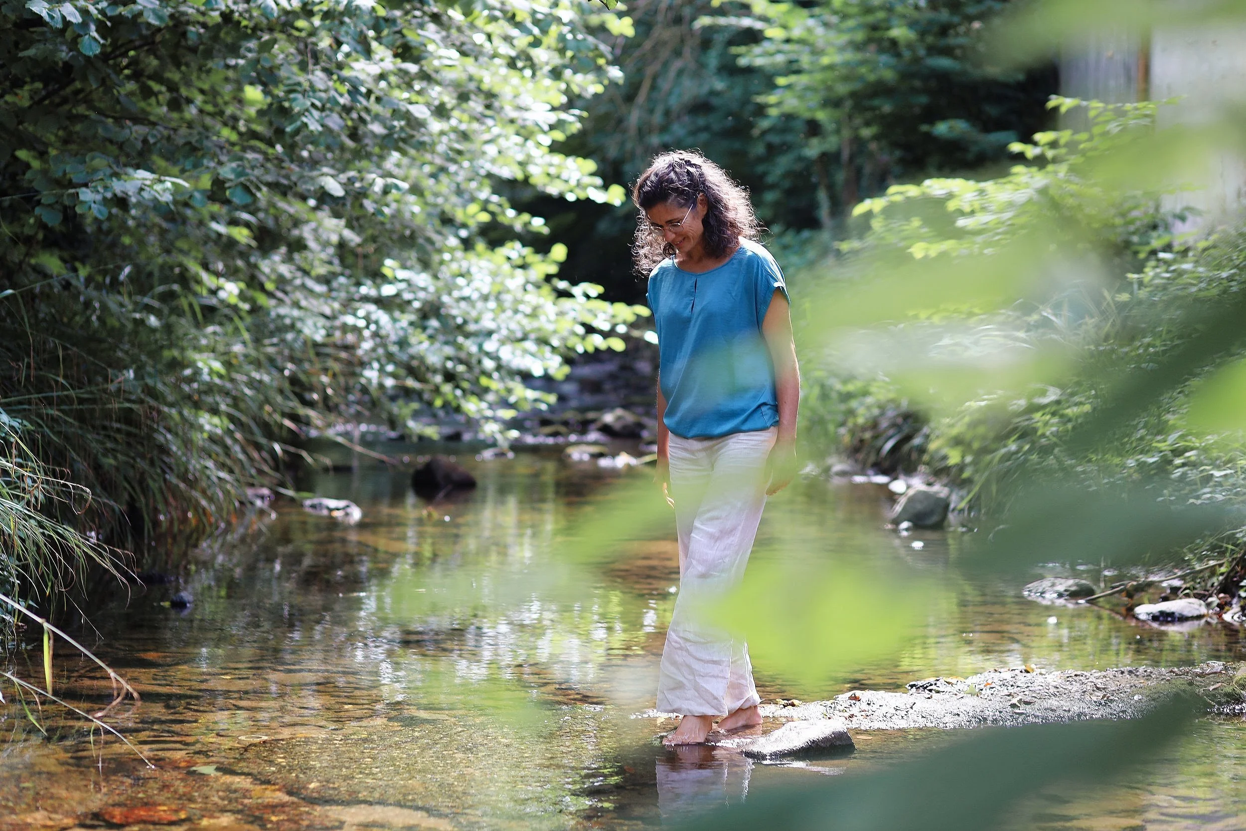 Frau in blauer Bluse und weißen Hosen steht barfuß in einem kleinen Fluss in einem grünen, bewaldeten Gebiet.