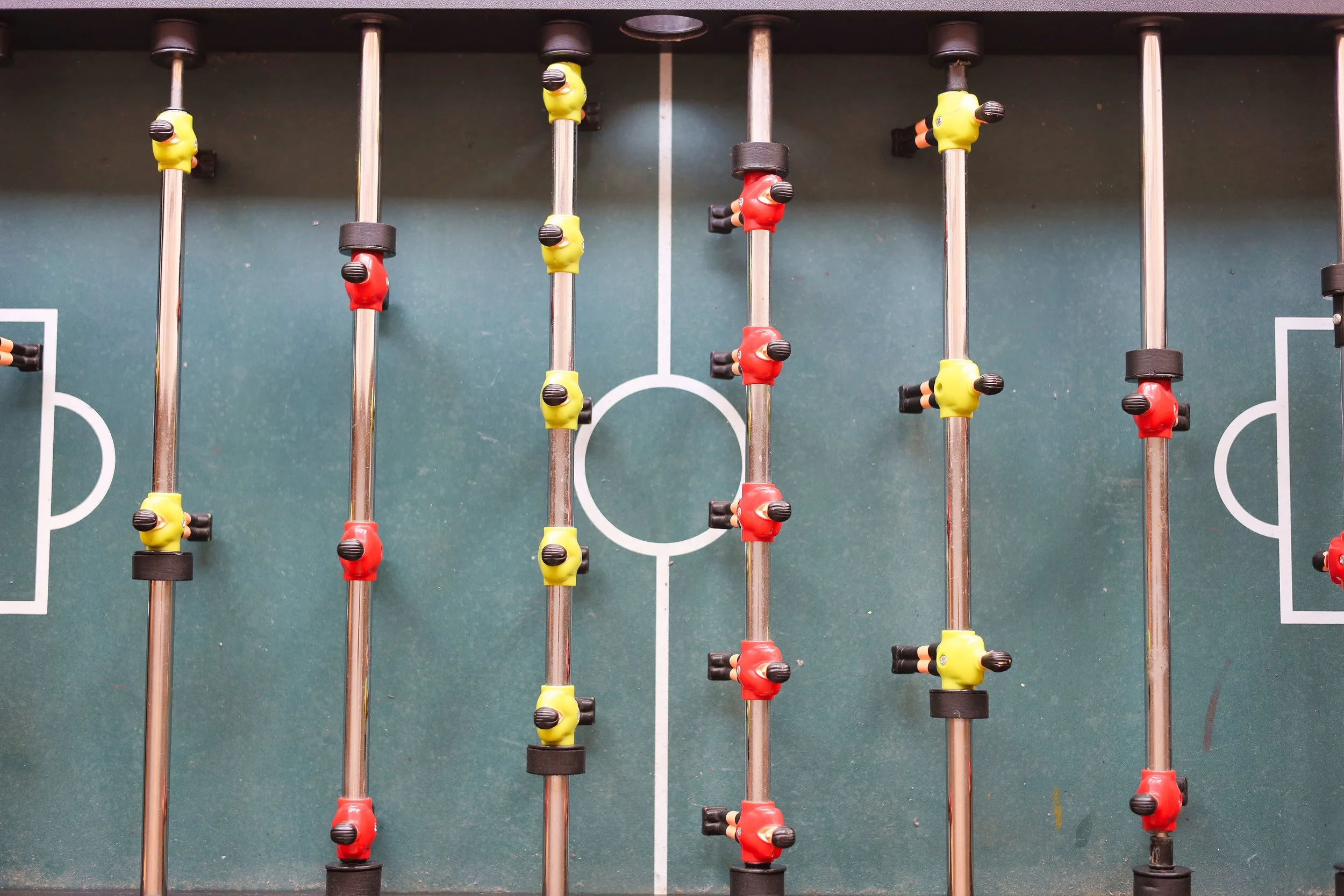 Fußballtisch mit grüner Spielfeldoberfläche und weißen Linien, Stangen mit roten und gelben Spielern, die in einem Foosball-Spiel angeordnet sind.