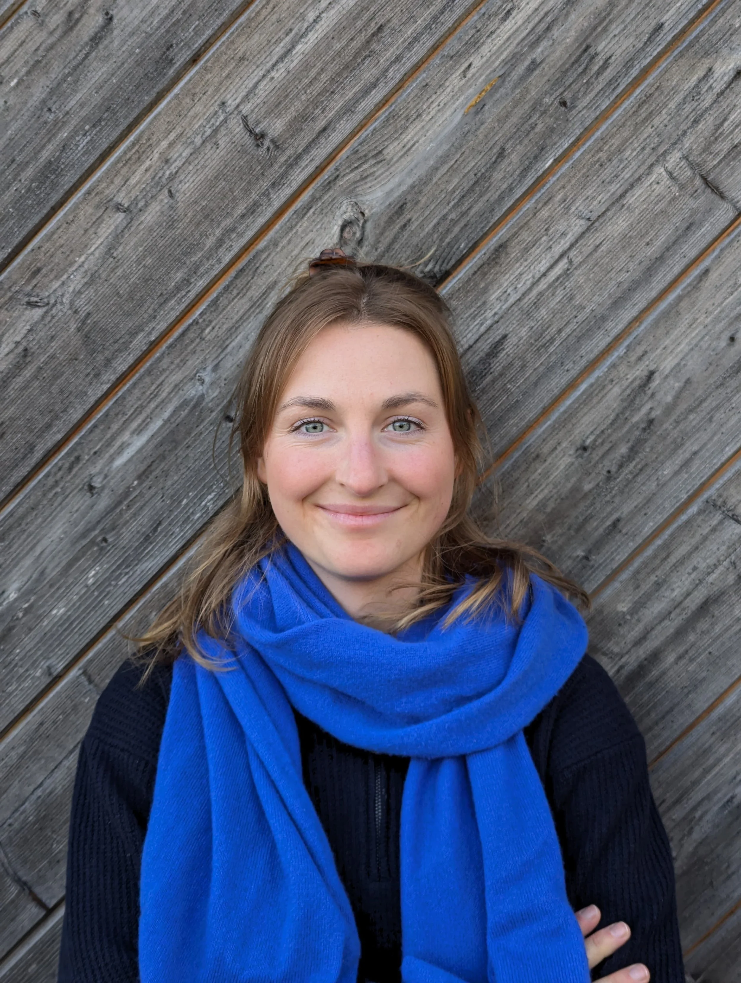 Eine lächelnde Frau mit braunen Haaren und blauen Augen vor einer Holzwand, trägt eine schwarze Jacke und einen blauen Schal.