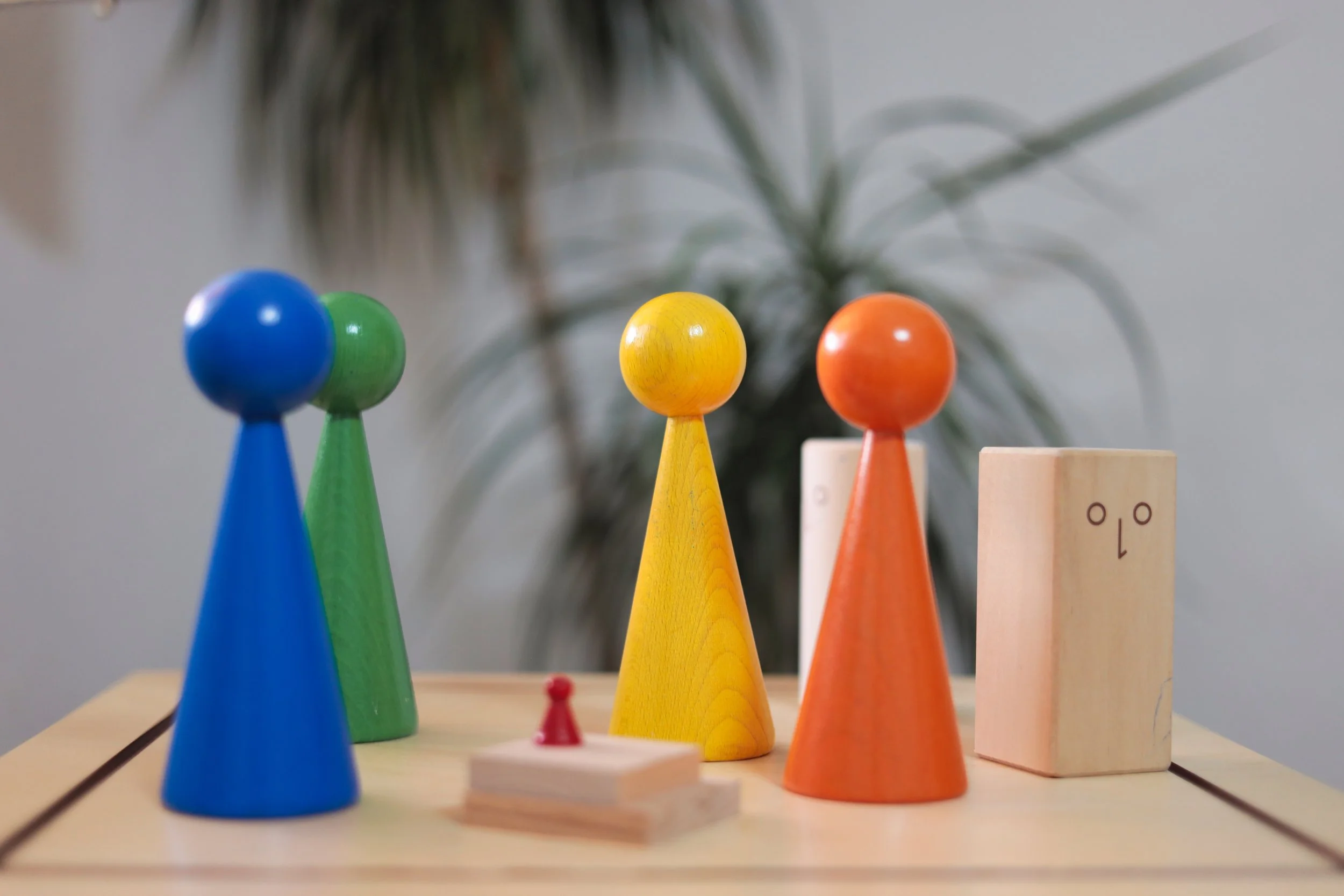 Bunte Holzspielfiguren in Blau, Grün, Gelb, Orange und einer hellen Holzfigur auf einem Holztisch, mit einer Pflanze im Hintergrund.