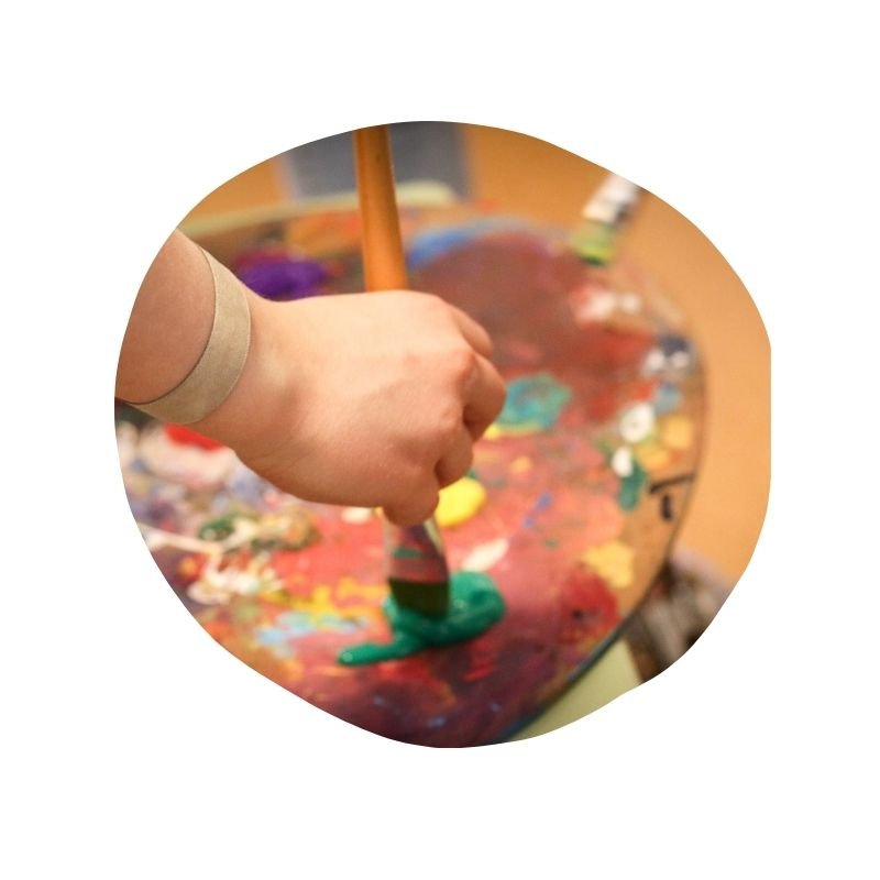 Nahaufnahme einer Hand, die mit bunten Farben auf einer Palette malt, umgeben von Farbklecksen.