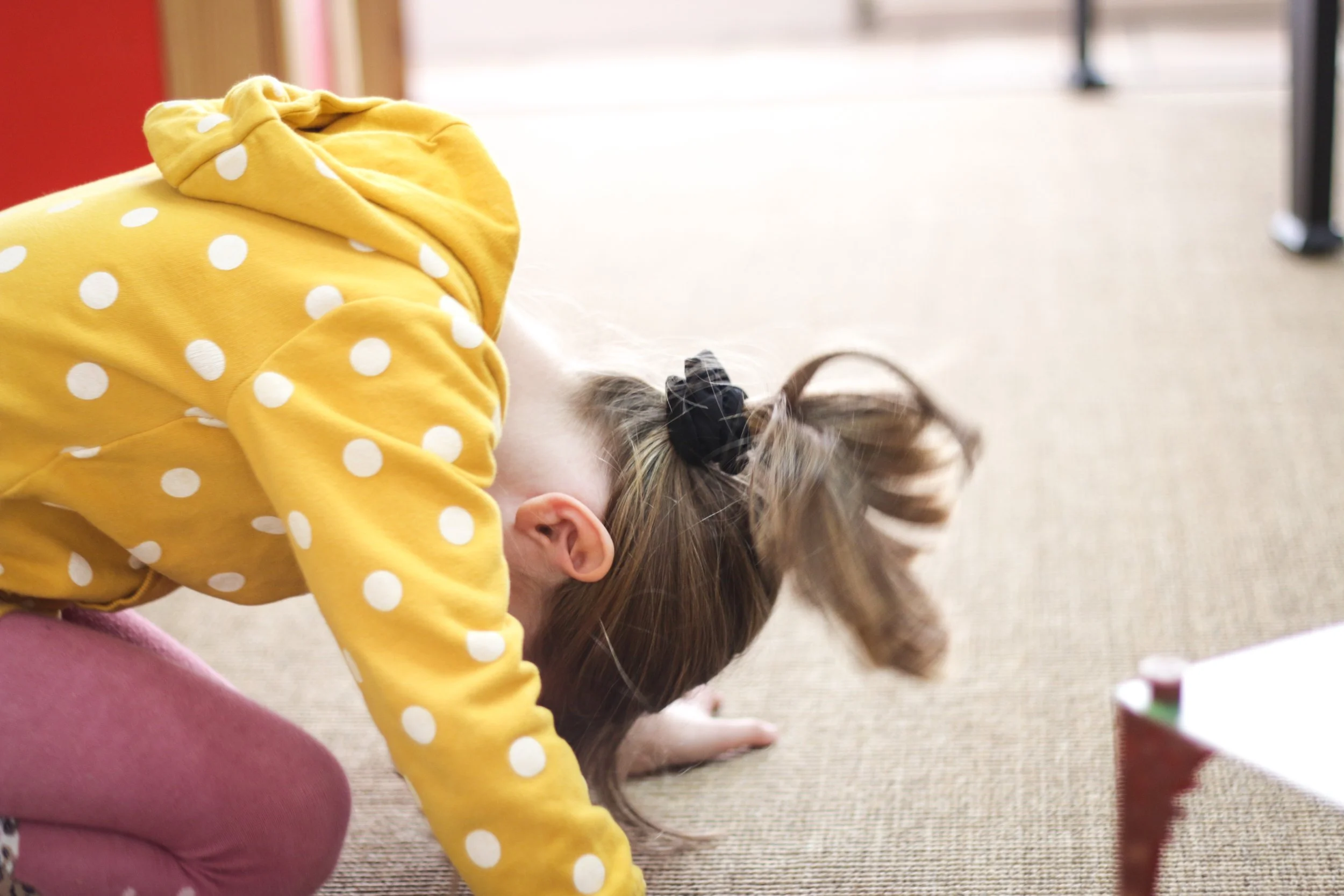 Ein Kind in einem gelben Pullover mit weißen Punkten und rosa Hosen, das auf dem Boden kniet und mit dem Kopf nach unten schaut.