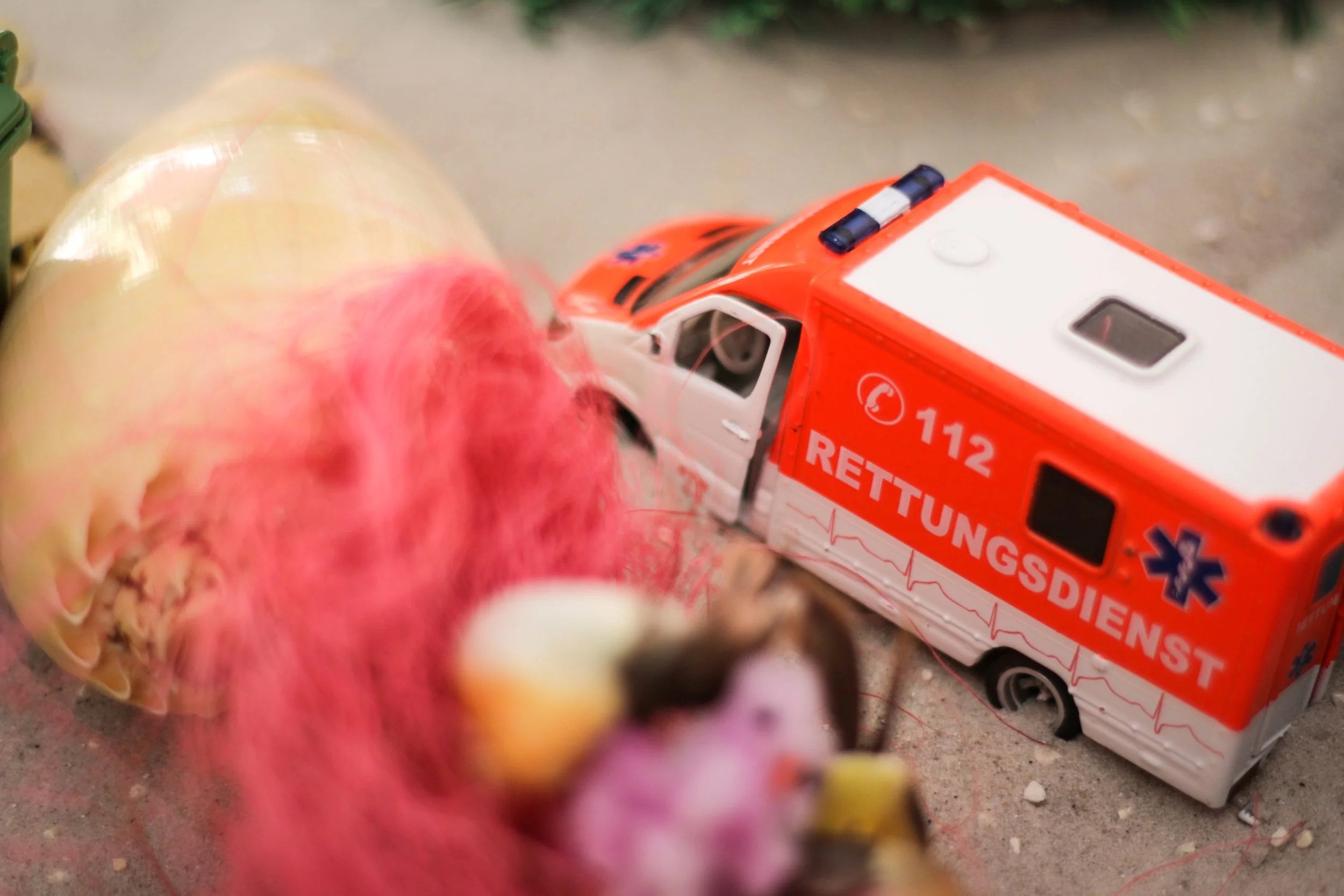 Modell eines Rettungswagens auf dem Boden, daneben ein pinkfarbenes und ein gelbes Objekt
