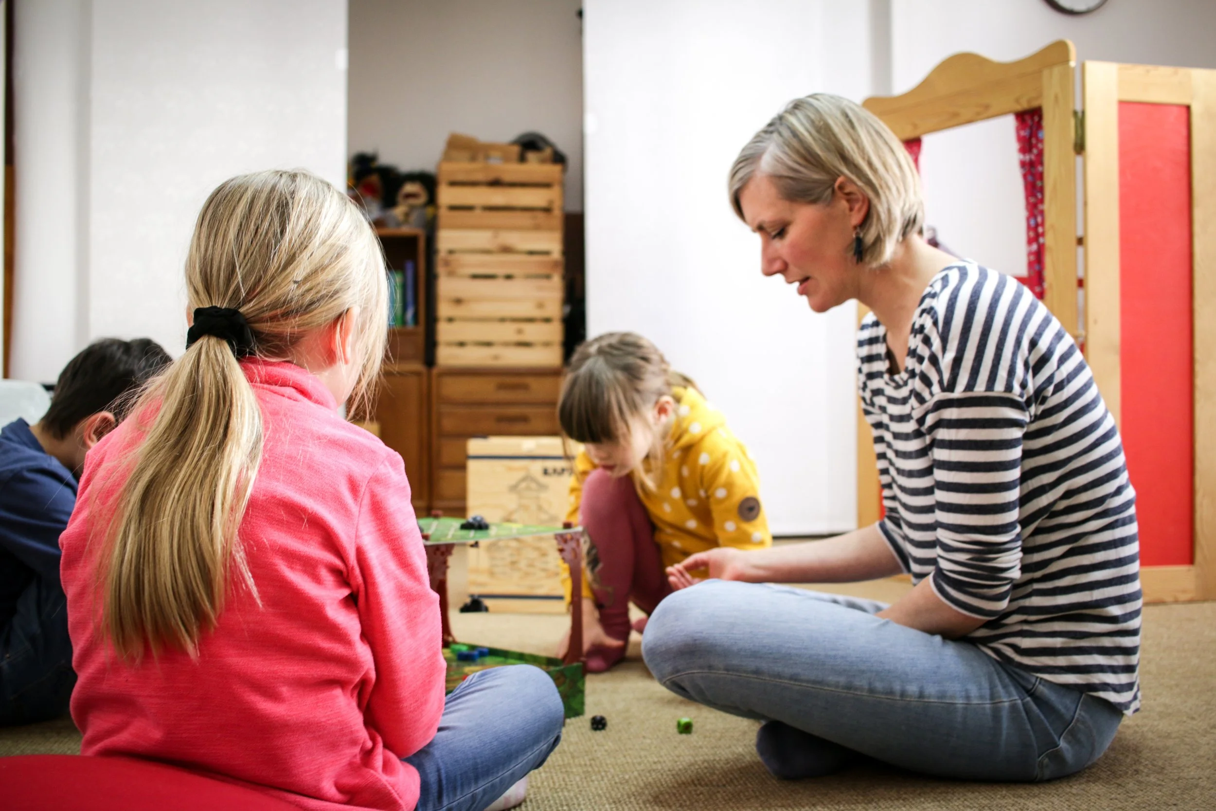 Eine Erwachsene spielt mit vier Kindern, die auf einem Teppich im Zimmer sitzen, während sie ein Brettspiel spielen.