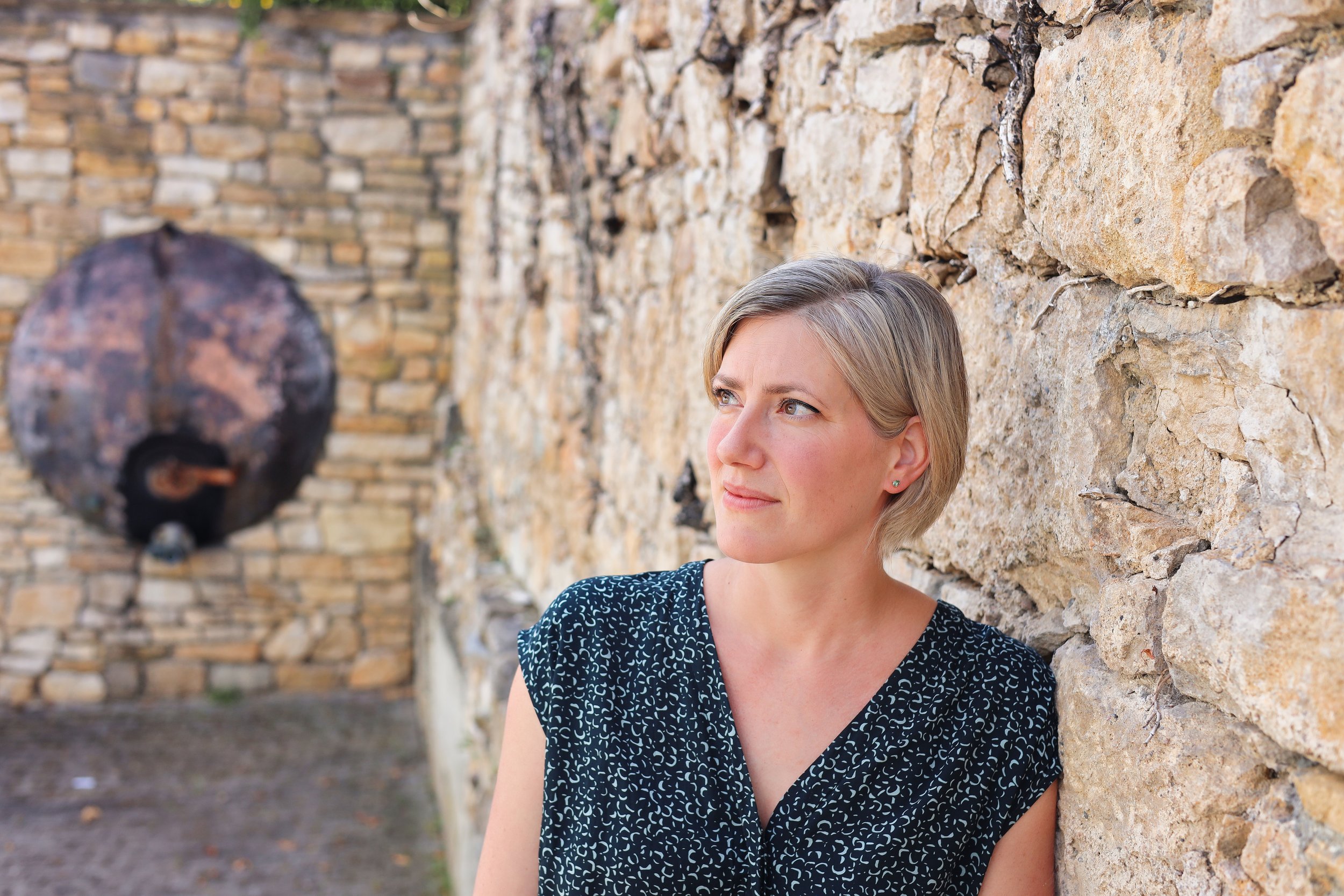 Junge Frau mit kurzen blonden Haaren steht an einer alten Steinmauer, blickt nach rechts und wirkt nachdenklich.
