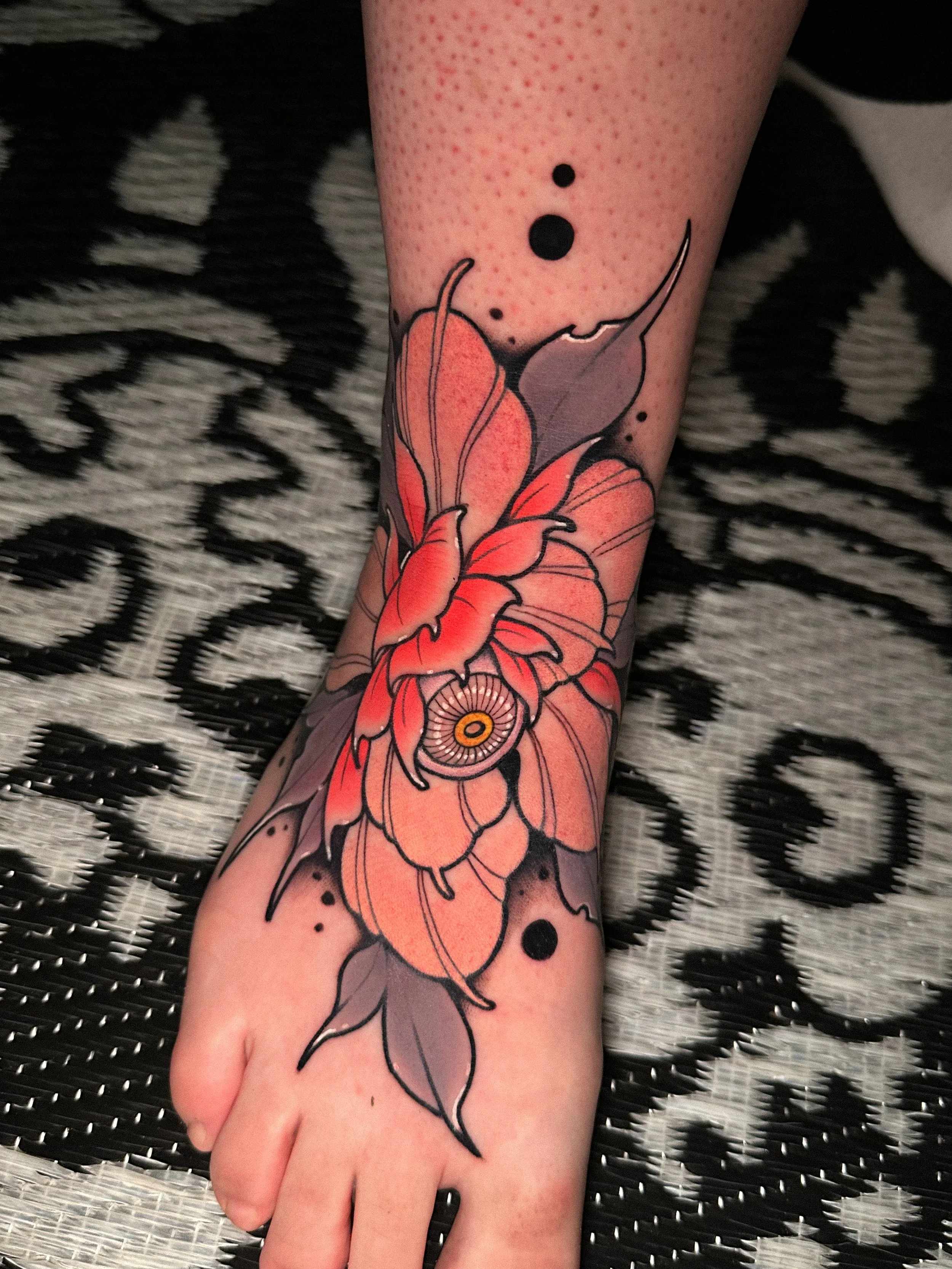 A person's forearm with a colorful floral tattoo design, featuring orange and purple flowers with black outlines and accents, resting on a black and white patterned surface.