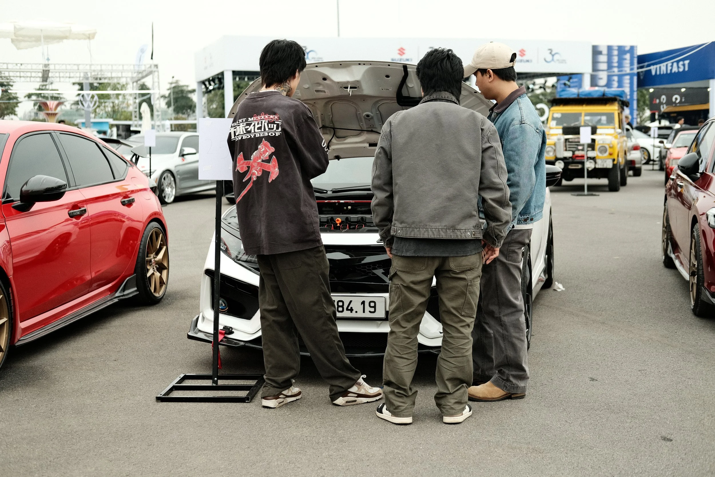Three young men stand in front of a white sports car with the hood open, examining the engine at an outdoor car show. There are other cars and people in the background.