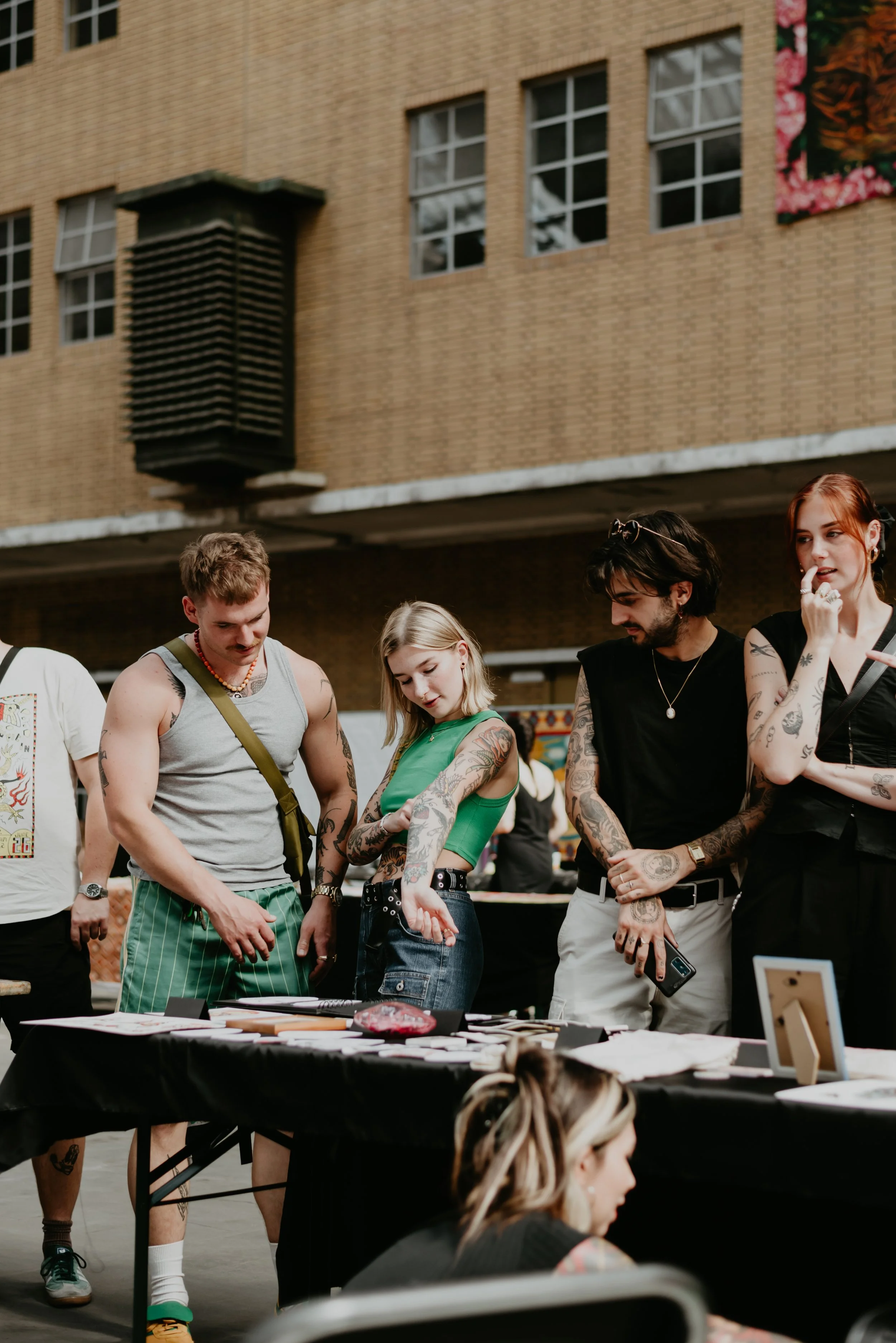 Group of young adults with tattoos standing around a table at an outdoor event, browsing items on display