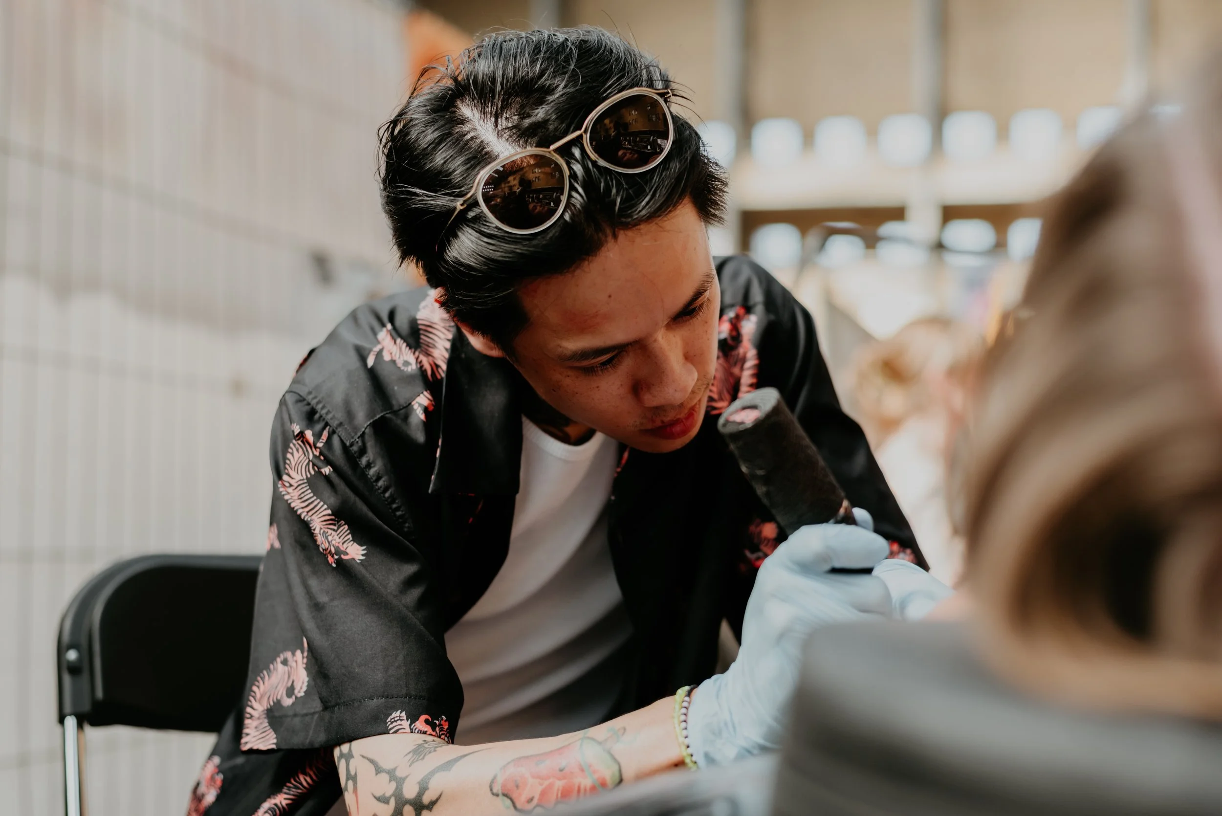 Person with dark hair, sunglasses on head, and tattoos on arms, getting a tattoo on their arm by a tattoo artist wearing gloves.