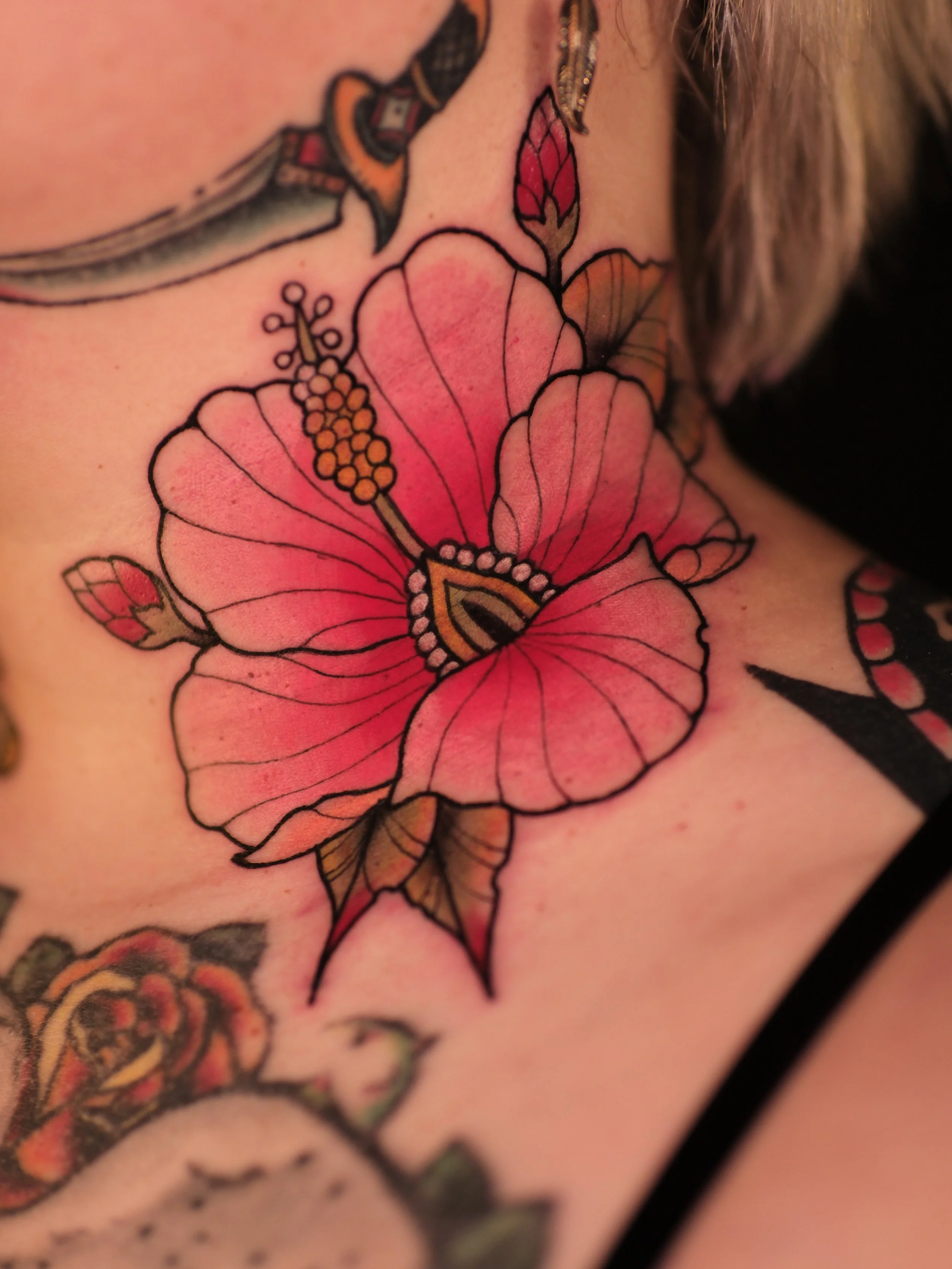Close-up of a colorful tattoo of a pink hibiscus flower, a red flower bud, and surrounding leaves on skin.