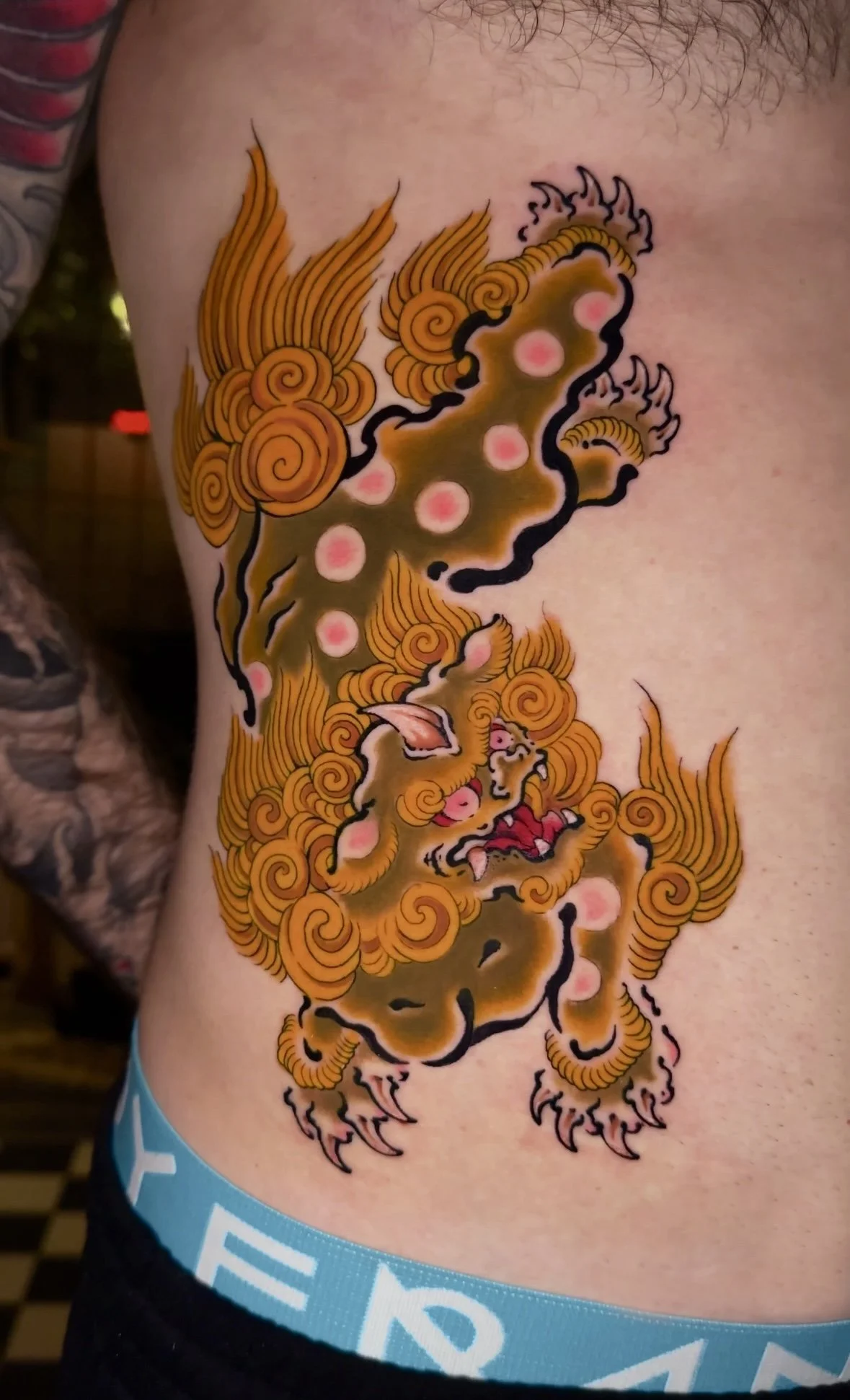Tattoo of a traditional Japanese dragon with orange and gold scales, black outlines, and pink accents on person's side.