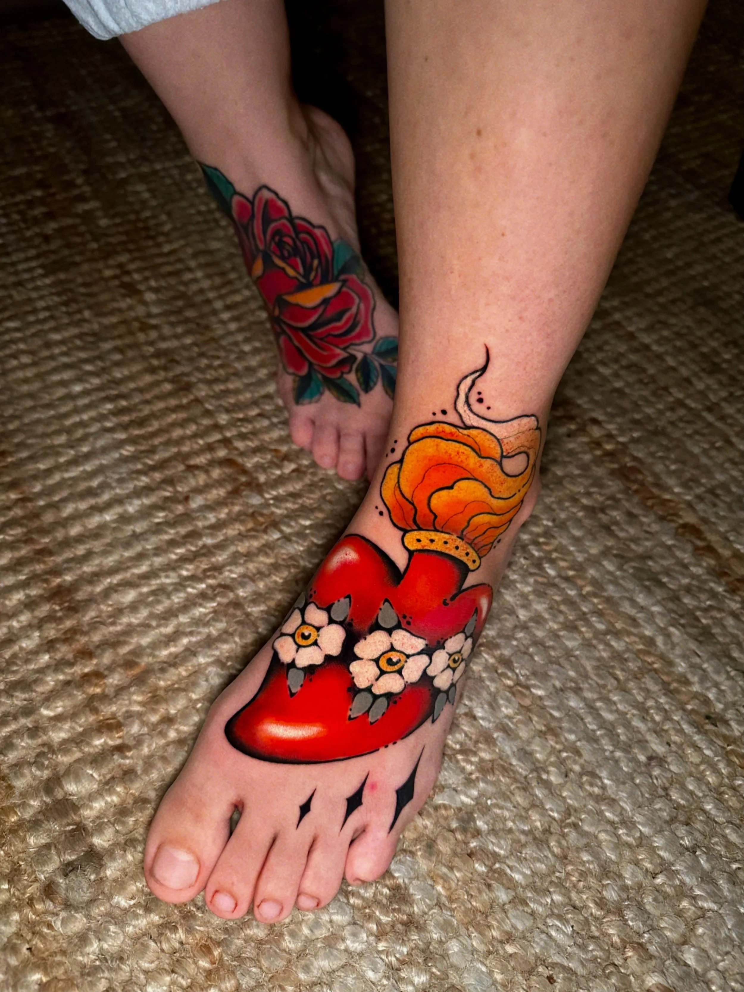 Close-up of a person's foot and lower leg with multiple colorful tattoos, including a red flower, orange flame, and white flowers, lying on a textured beige carpet.