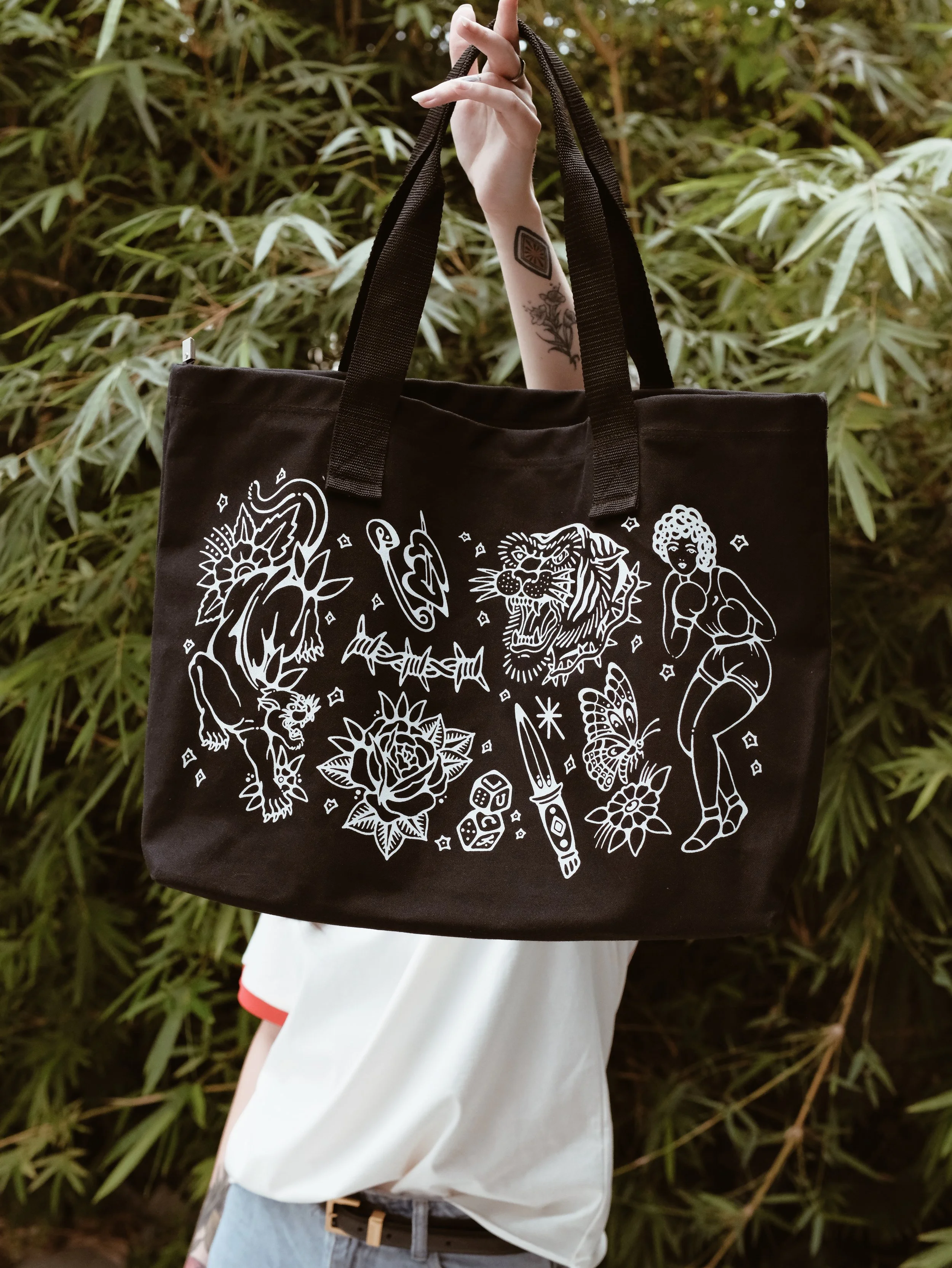 A person holding a black tote bag with white tattoo-style illustrations of animals, a woman, and various objects, outdoors with green foliage in the background.