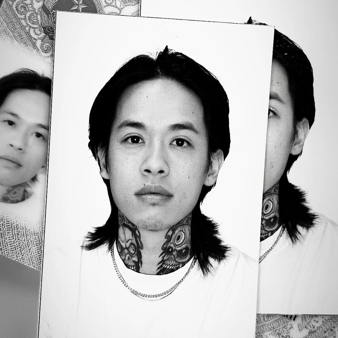 A black-and-white portrait of a young person with shoulder-length hair, a tattoo on their neck, and two necklaces, looking directly at the camera.