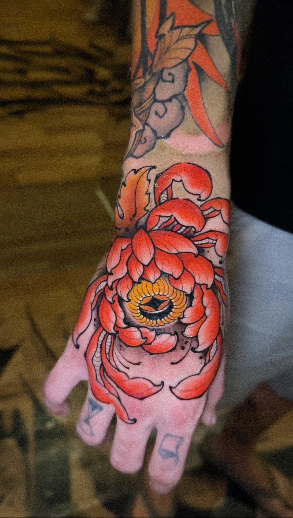 A person's arm and hand with a colorful tattoo sleeve featuring a red and orange flower, leaves, and abstract design.