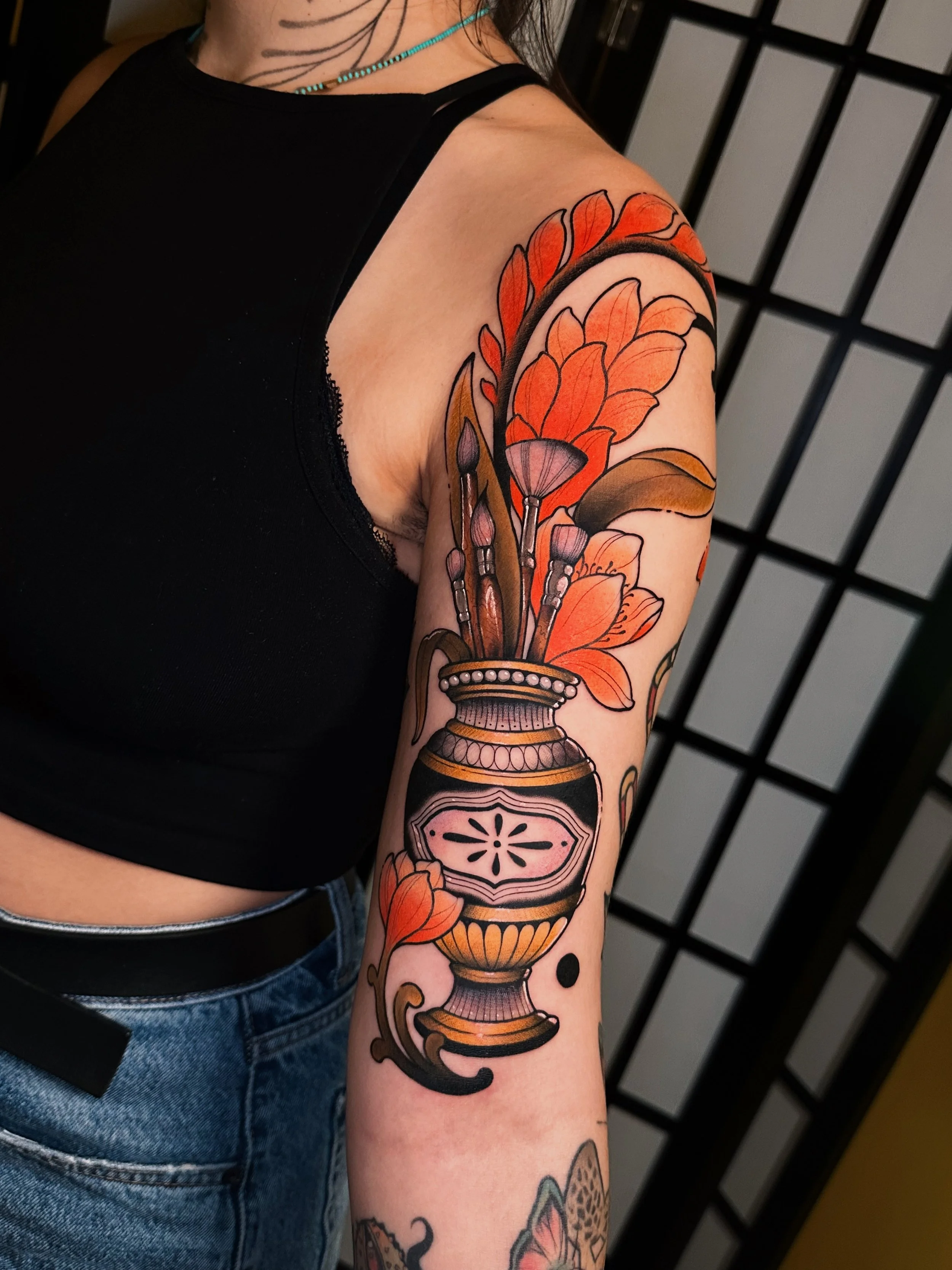 A person's upper arm tattoo featuring orange and pink flowers in a vase with black outlines, clay-like details, and several paintbrushes.