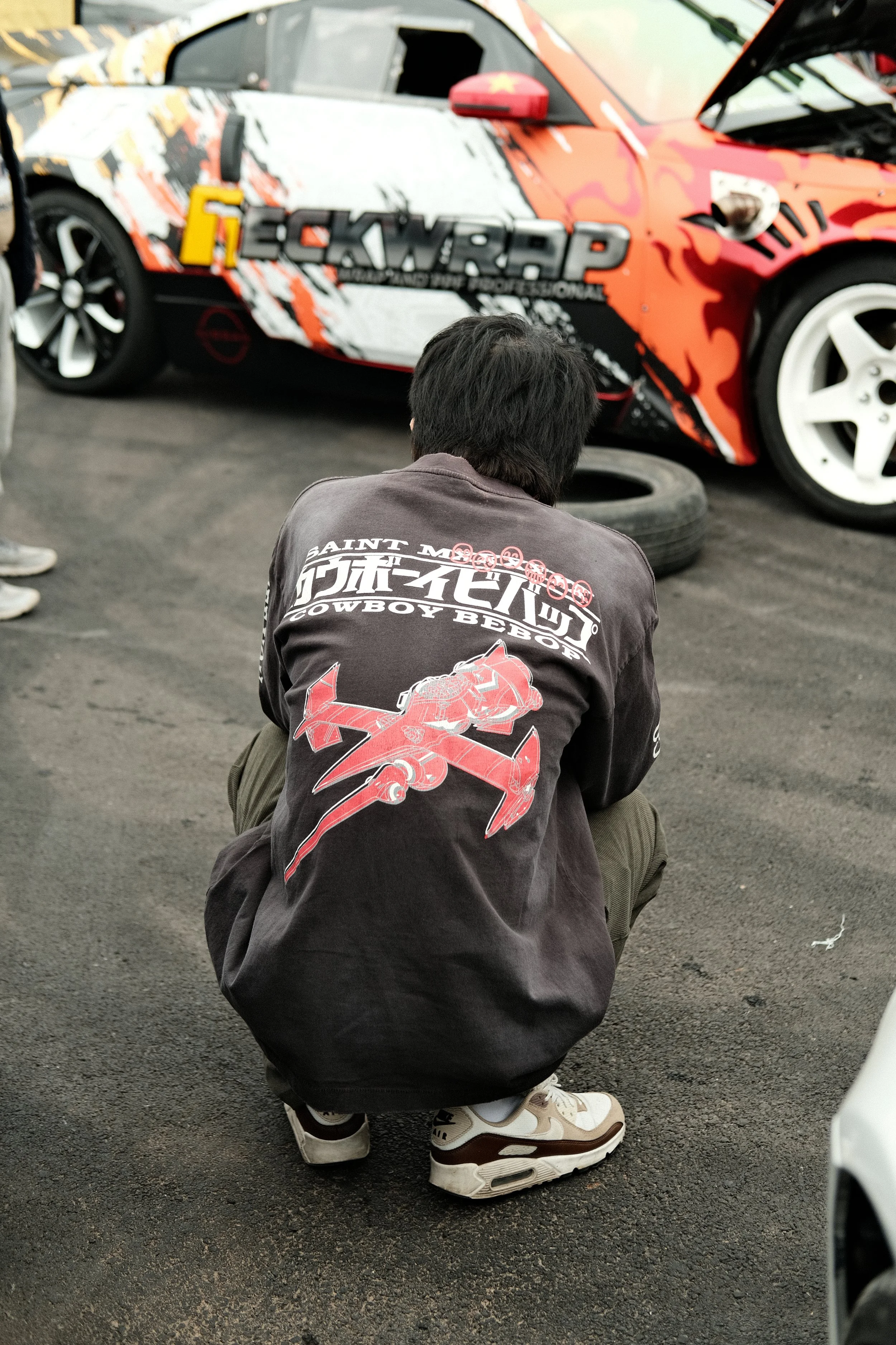 A person crouching on the pavement in front of a race car with a colorful, graphic design. The person is wearing a dark sweatshirt, beige sneakers, and dark pants.