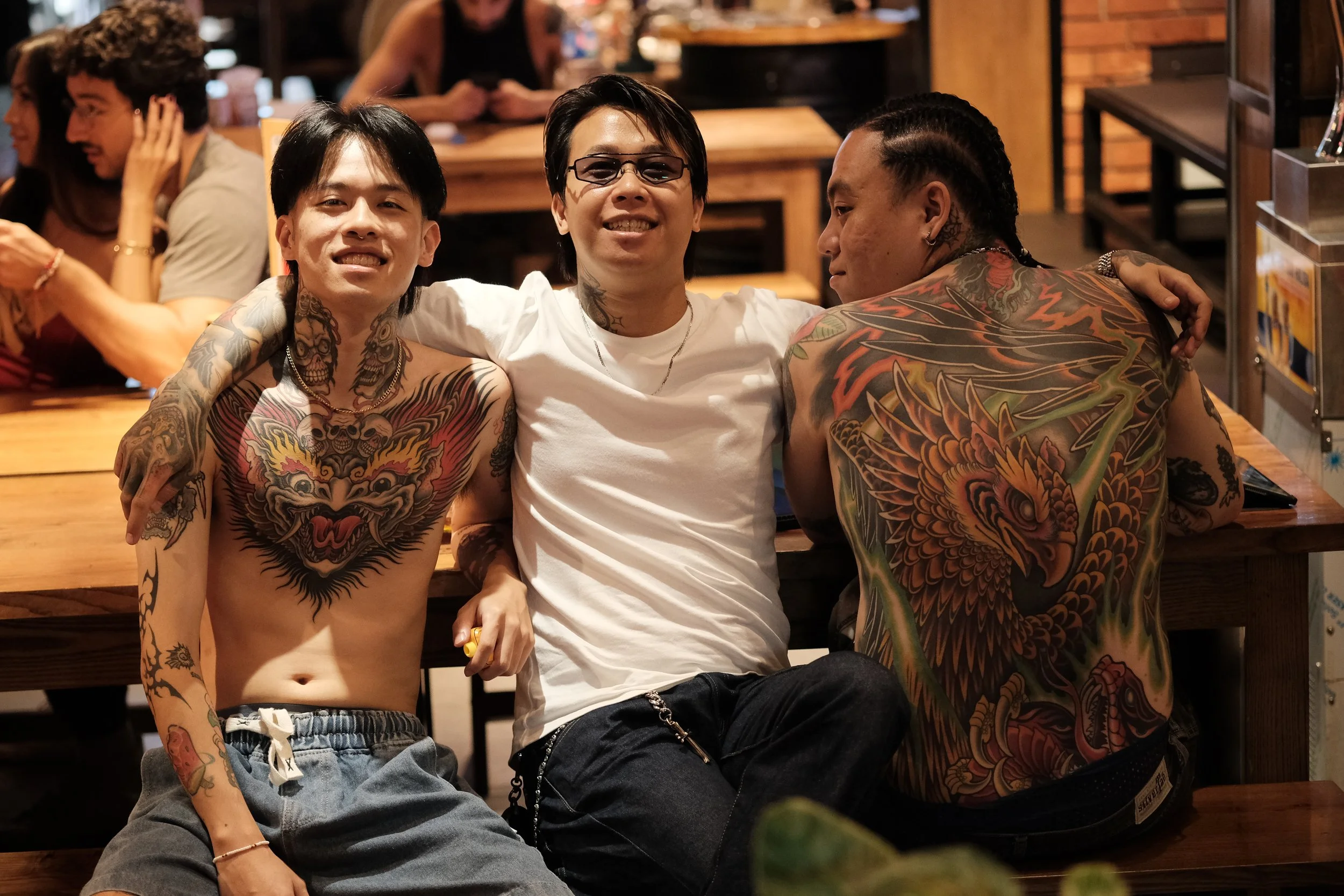 Three young men with colorful tattoos sitting together in a restaurant, smiling at the camera, with other customers in the background.