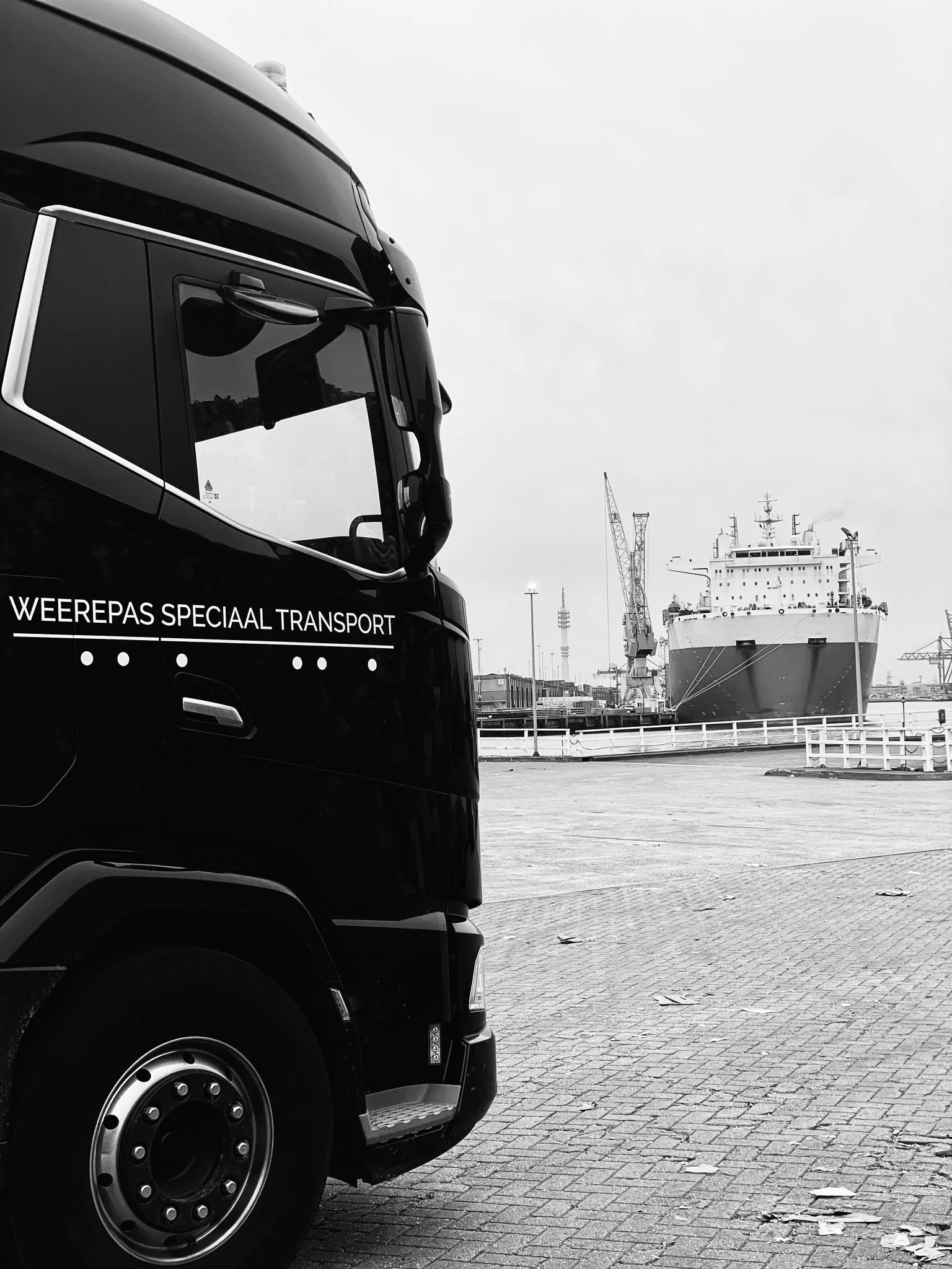 Close-up van het voorgedeelte van een zwart vrachtwagen met de tekst 'WEEREPAS SPECIAAL TRANSPORT' op de zijkant, geparkeerd op een haven met een schip en scheepskranen op de achtergrond.