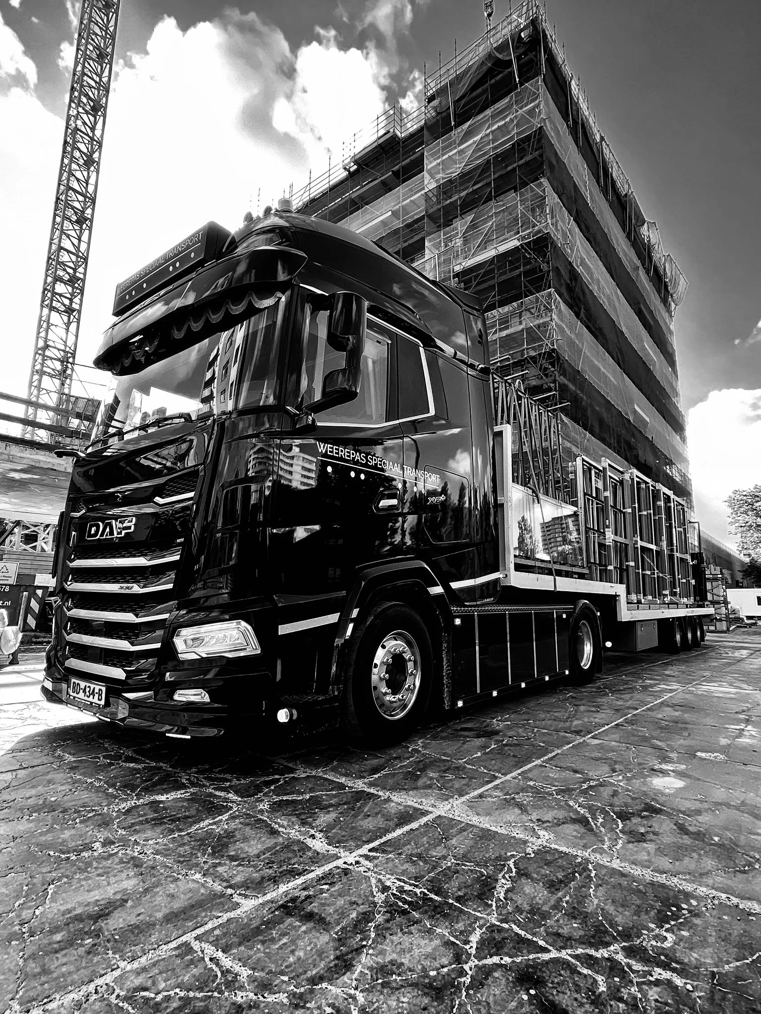 Zwart vrachtwagen met bouwmaterialen geladen, geparkeerd op een bestrate markt, met een bouwproject in de achtergrond en een torenkraan.