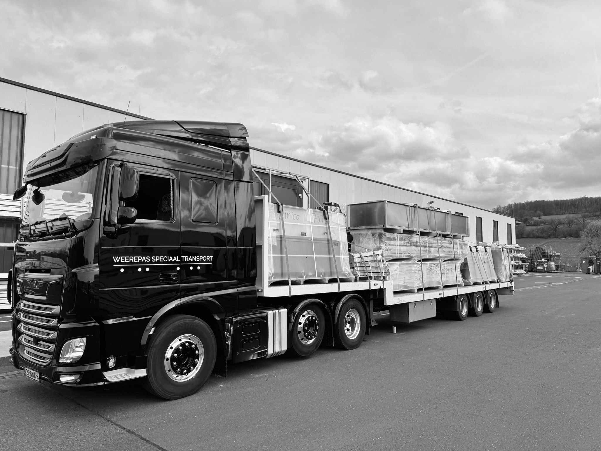 Zwart vrachtwagen met de tekst 'WEEREPAS SPECIAAL TRANSPORT' geladen met grote panelen, geparkeerd buiten voor een industrieel gebouw, onder een bewolkte hemel.