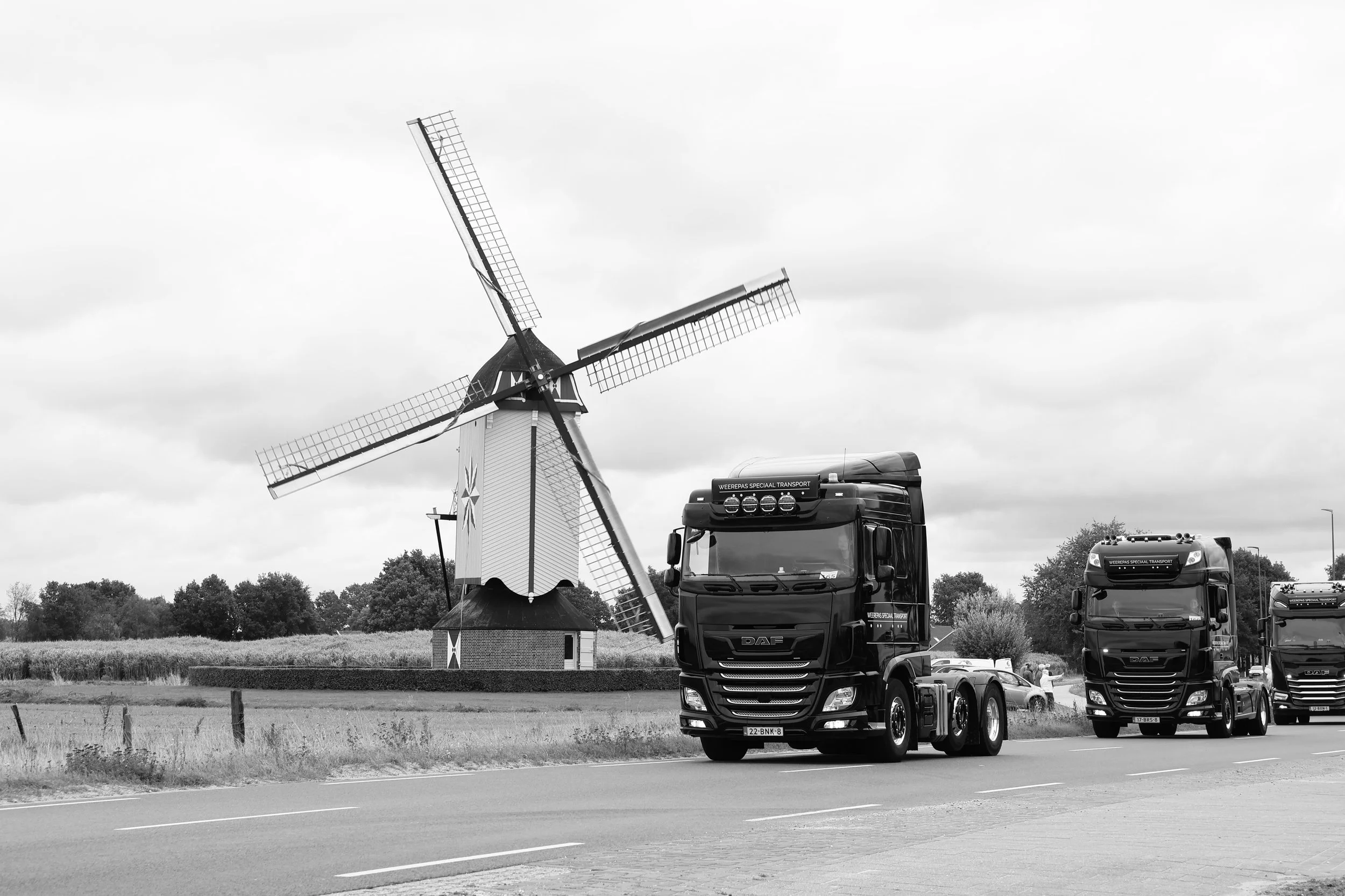 Een zwart-wit afbeelding van een molen met draaiende wieken in een open landschap, met enkele vrachtwagens op de weg eronder.