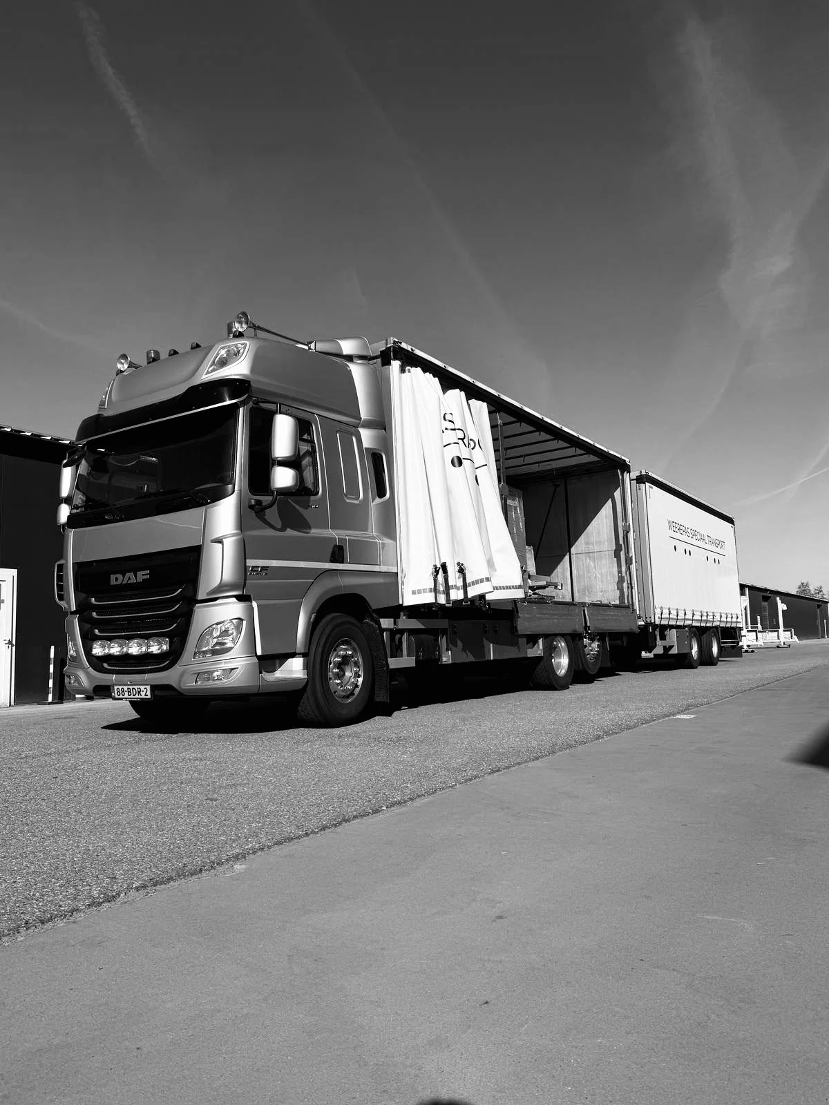 Zwart-witte foto van een grote vrachtwagen met een open laadruimte langs een weg, onder een heldere hemel.