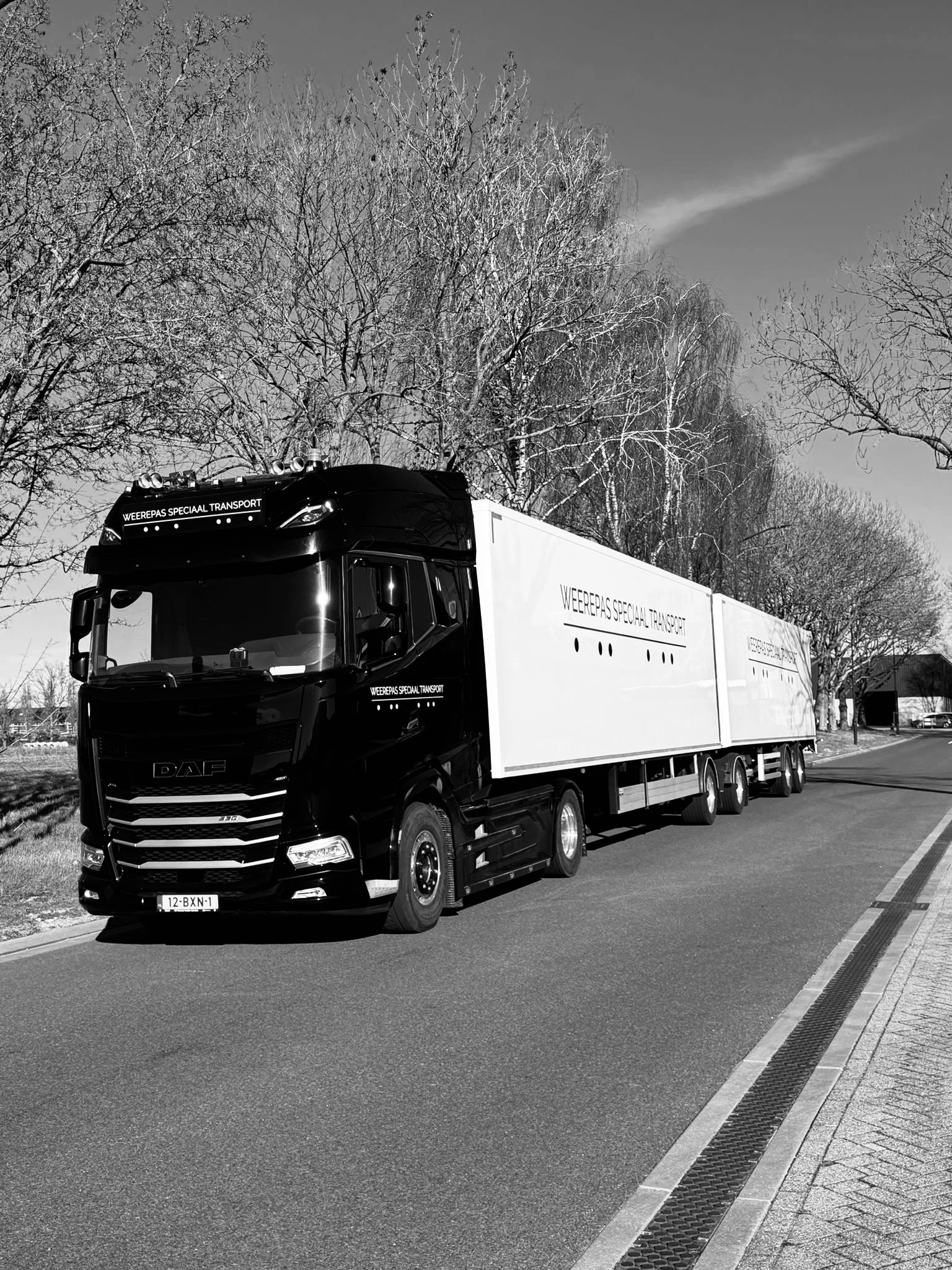 Zwarte vrachtwagen met witte trailer, rijdend op een weg, met kale bomen en een blauwe hemel op de achtergrond.