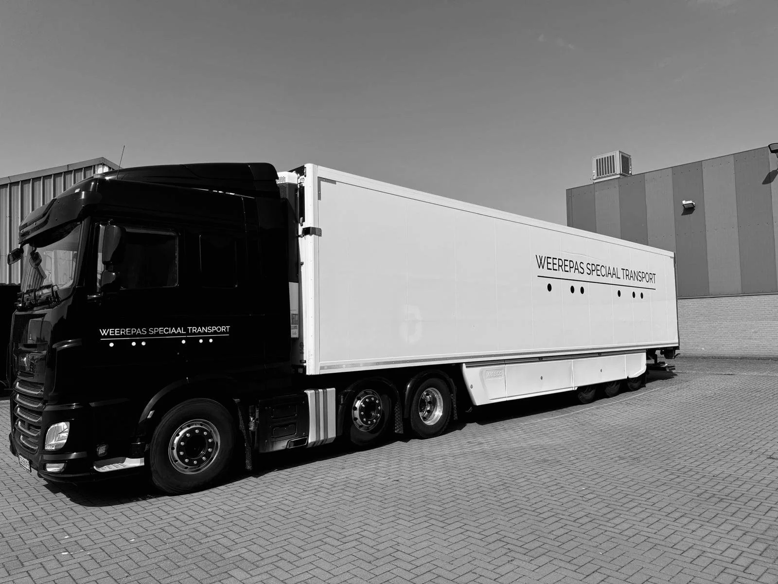 Zwarte vrachtwagen met witte container, gestationeerd op een parkeerplaats, met tekst 'Weerepas speciaal transport' op beide zijden van de container.