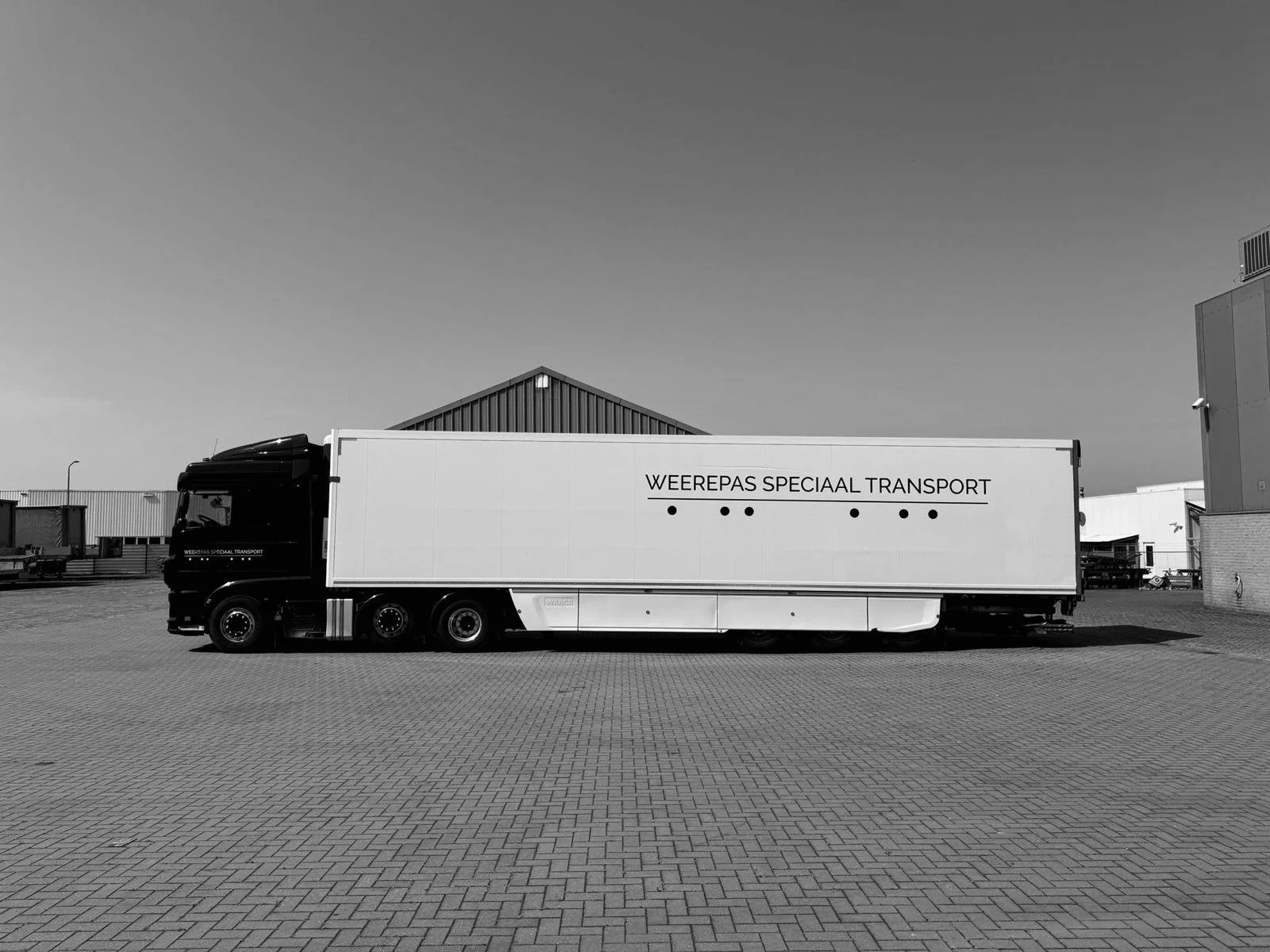 Zwart-witte foto van een vrachtwagen met de tekst "WEEREPAS SPECIAAL TRANSPORT" op de zijkant, geparkeerd op een betegelde ruimte voor een loods.