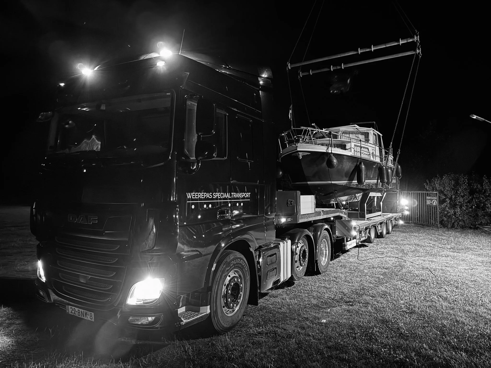 Een zwart-witte foto van een grote vrachtwagen die een boot vervoert, geparkeerd op een grasveld 's nachts, met duidelijke koplampen en een donkere achtergrond.