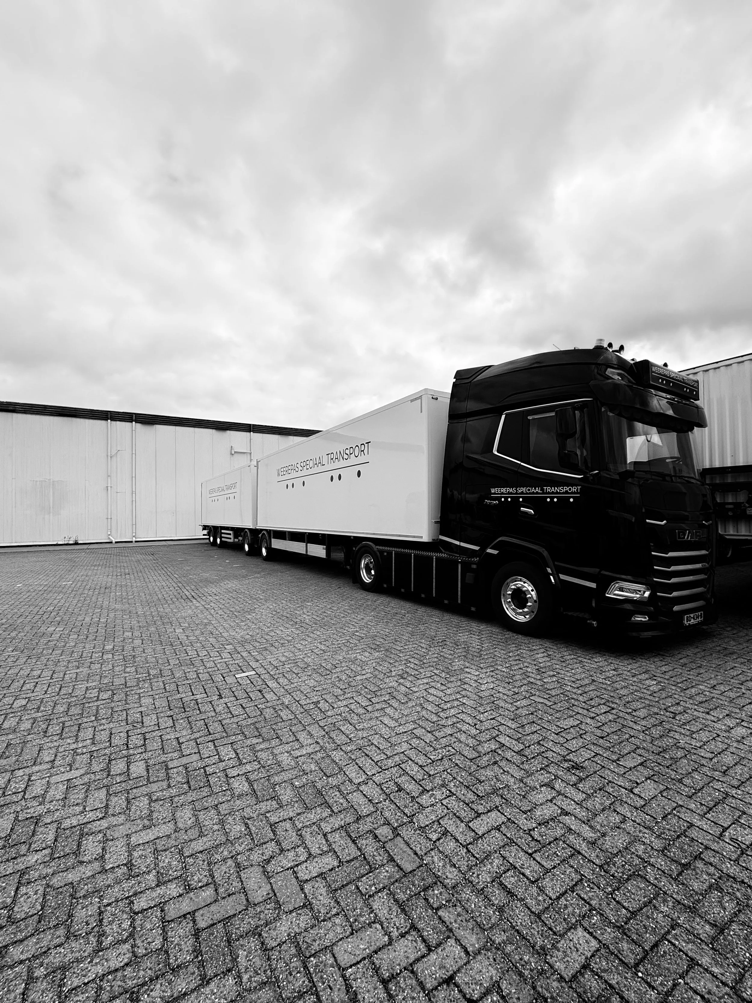 Een zwart truck met witte laadbak staat geparkeerd op een betegeld terrein bij een industrieel gebouw, onder een bewolkte hemel.