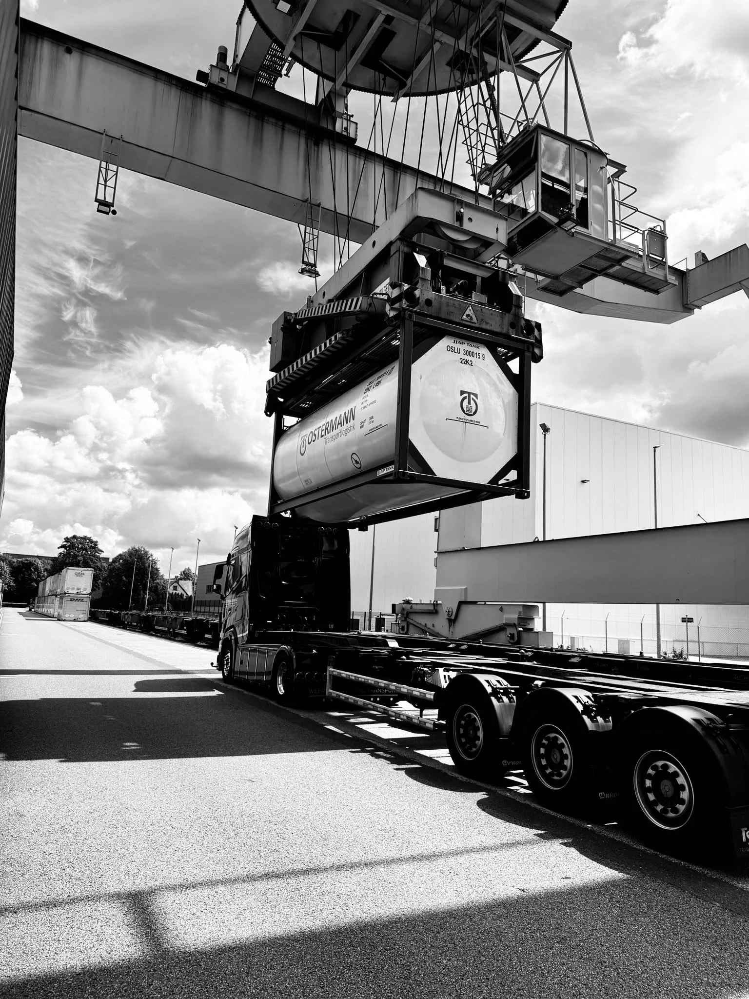 Een vrachtwagen laadt een grote container op een containertransportwagen, ondersteund door een brugkraan. De omgeving lijkt een logistiek faciliteit met wolken en gebouwen op de achtergrond.