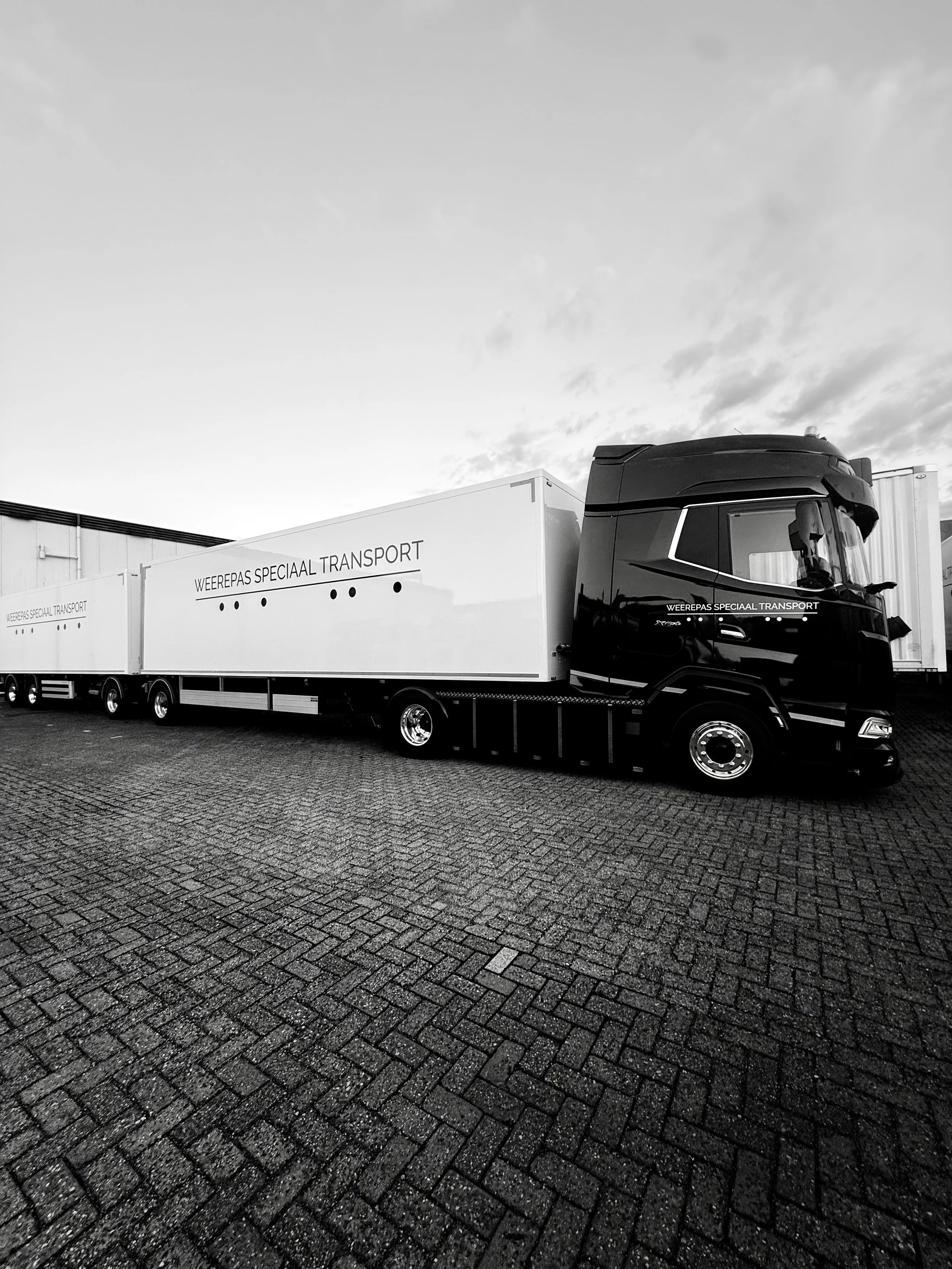 Zwart-witte vrachtwagen met de tekst 'Weerepas speciaal transport' geparkeerd op een stenen parkeerplaats onder een bewolkte hemel.