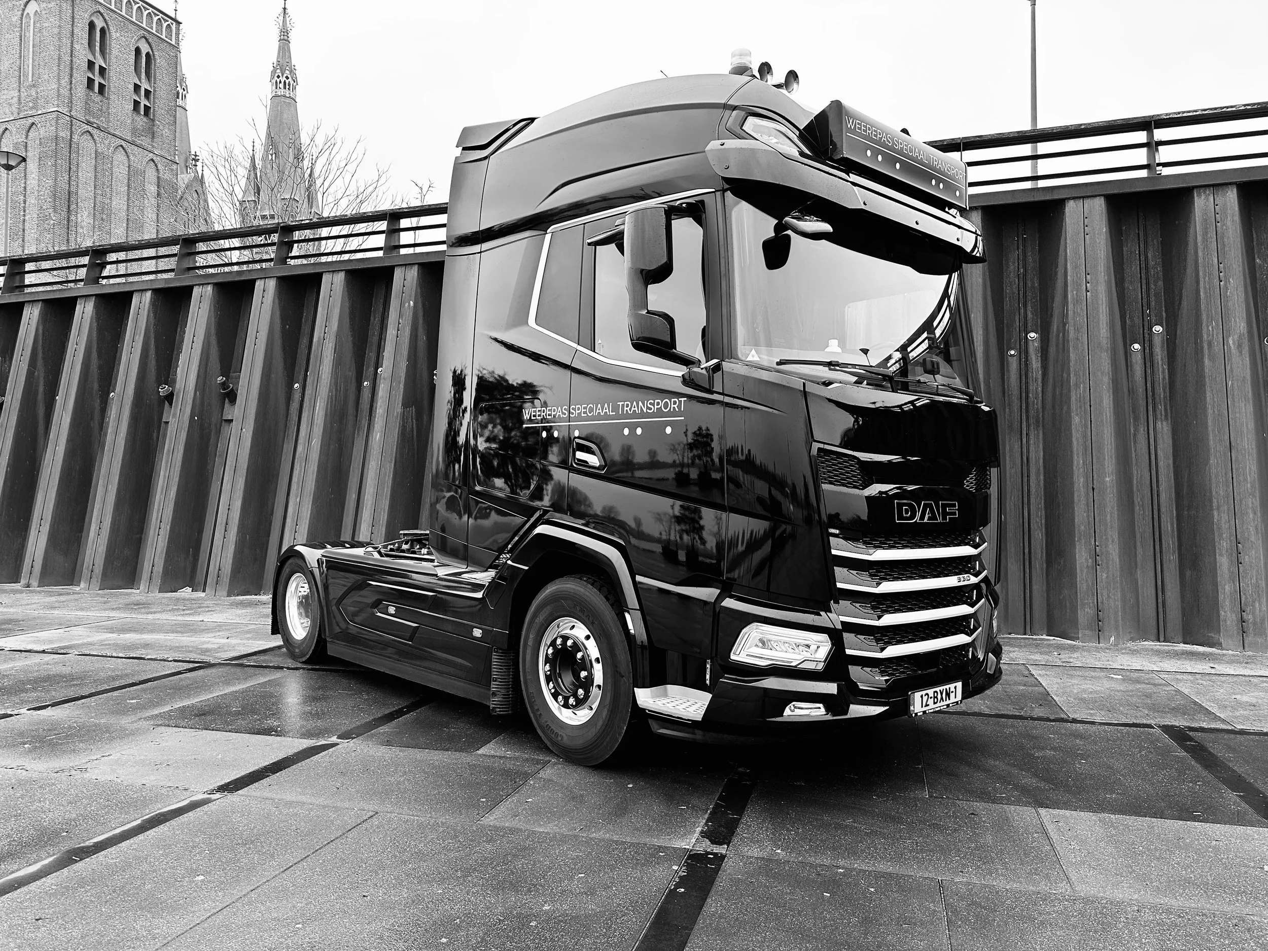 Zwart-truck met de tekst 'Weerepas speciaal transport' op de voorkant, geparkeerd naast een houten muur en een kerk met torens op de achtergrond, in een parking op een vochtige dag.