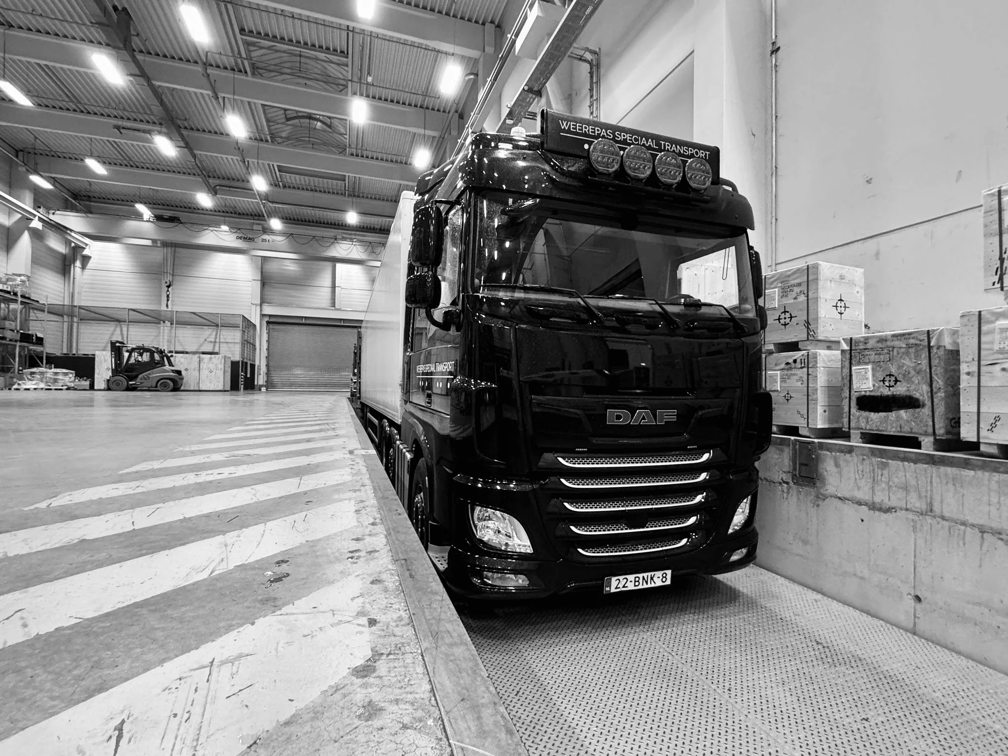 Een zwart truck met een een witte laadbak geparkeerd in een magazijn of opslagruimte, omgeven door pallets en dozen. De voorkant van de truck is prominent in beeld, met een nummerbord zichtbaar onderaan.