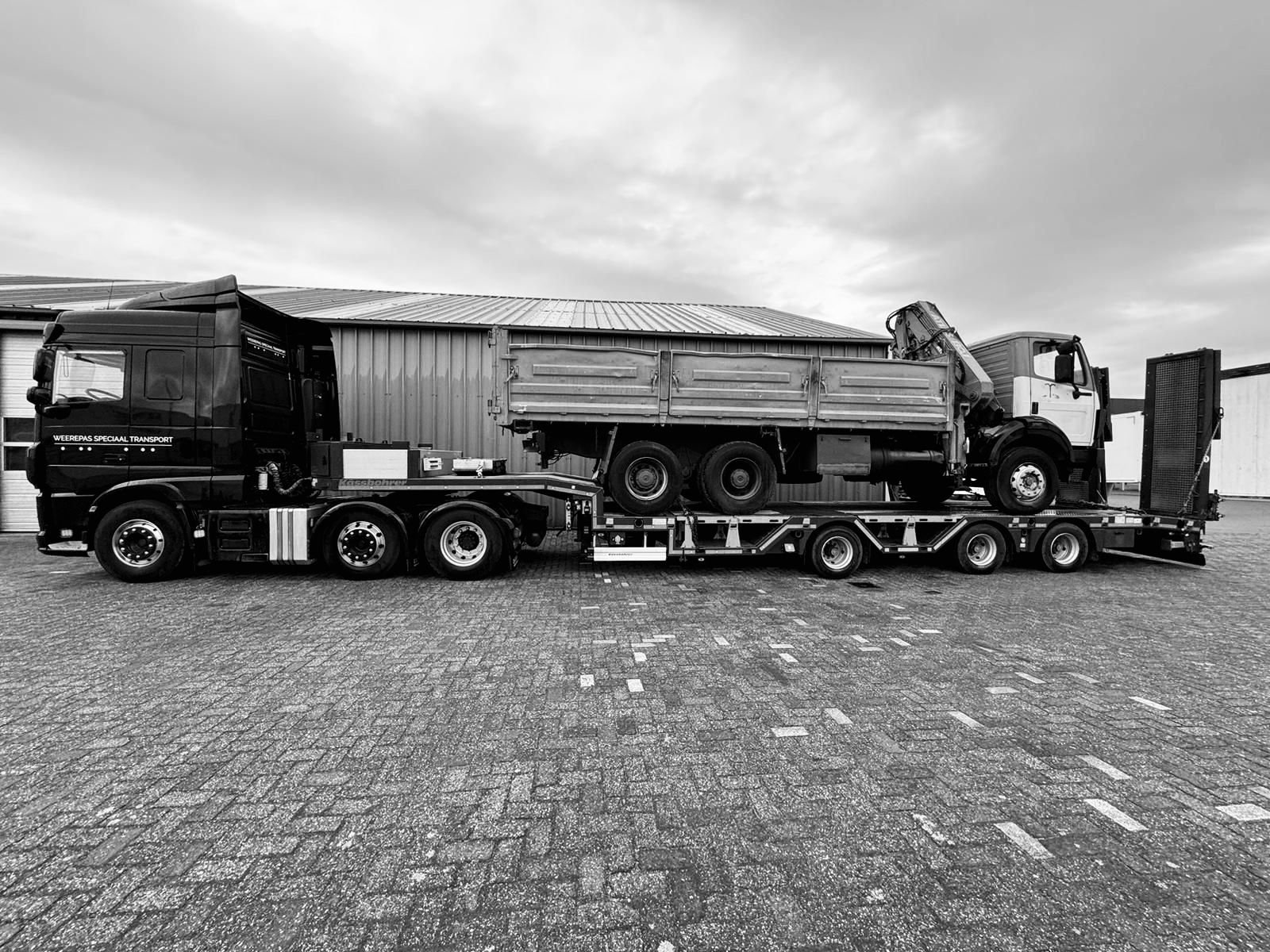 Een vrachtwagen op een dieplader, klaar om te reizen, geparkeerd op een bestraten oppervlak met een industrieel gebouw op de achtergrond.