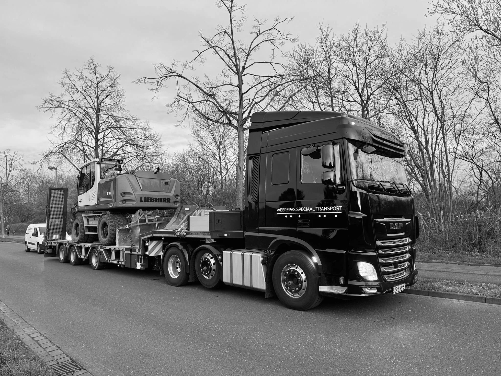Een grote vrachtwagen met een auto en een bouwmachine op de laadklep, geparkeerd langs een weg met bomen zonder bladeren op de achtergrond.