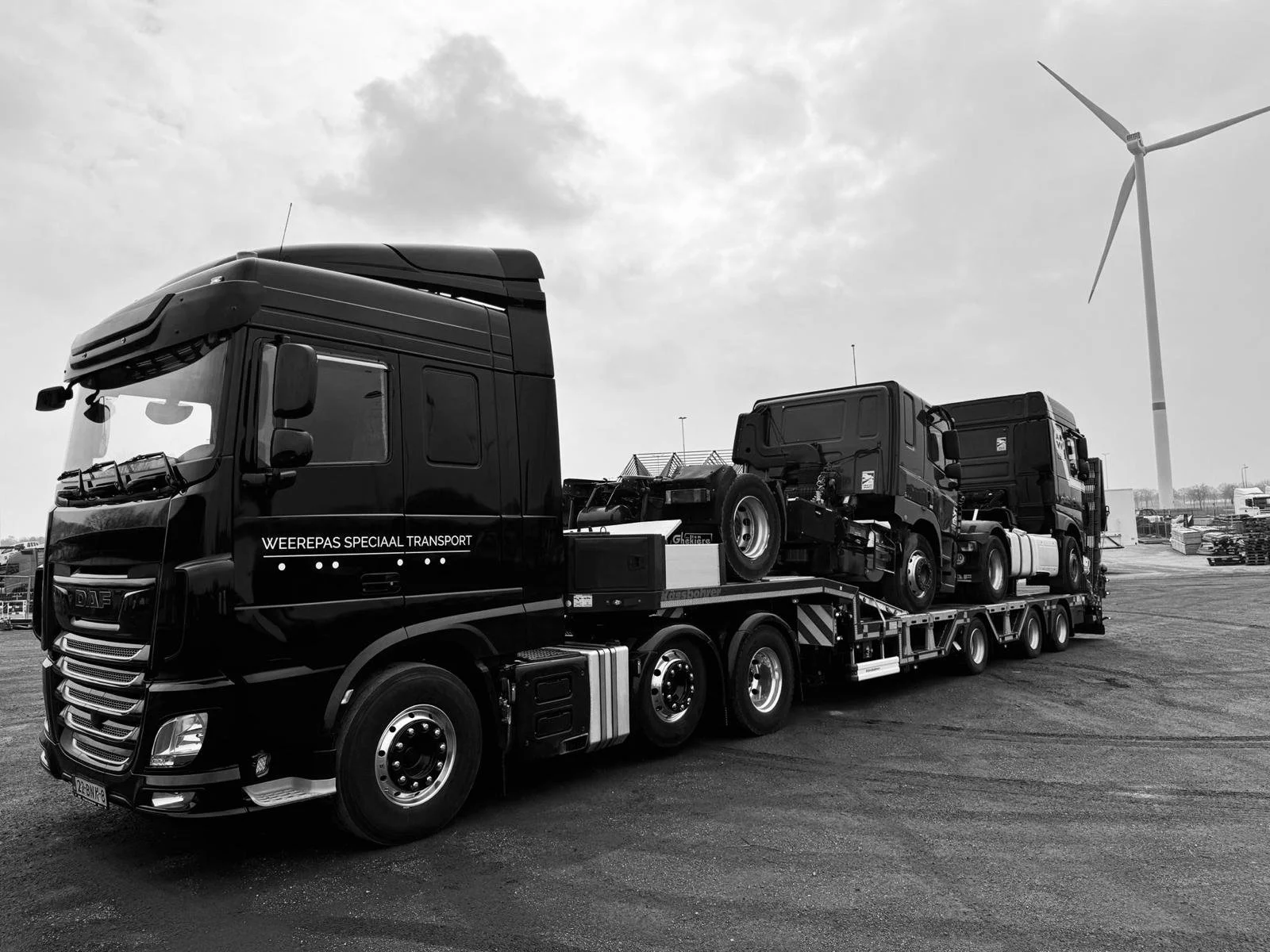 Zwart treintruck met transportwagen en twee vrachtwagens, één met opzetstuk, op een weg met windmolen en industrie op de achtergrond, in zwart-wil kleuren.