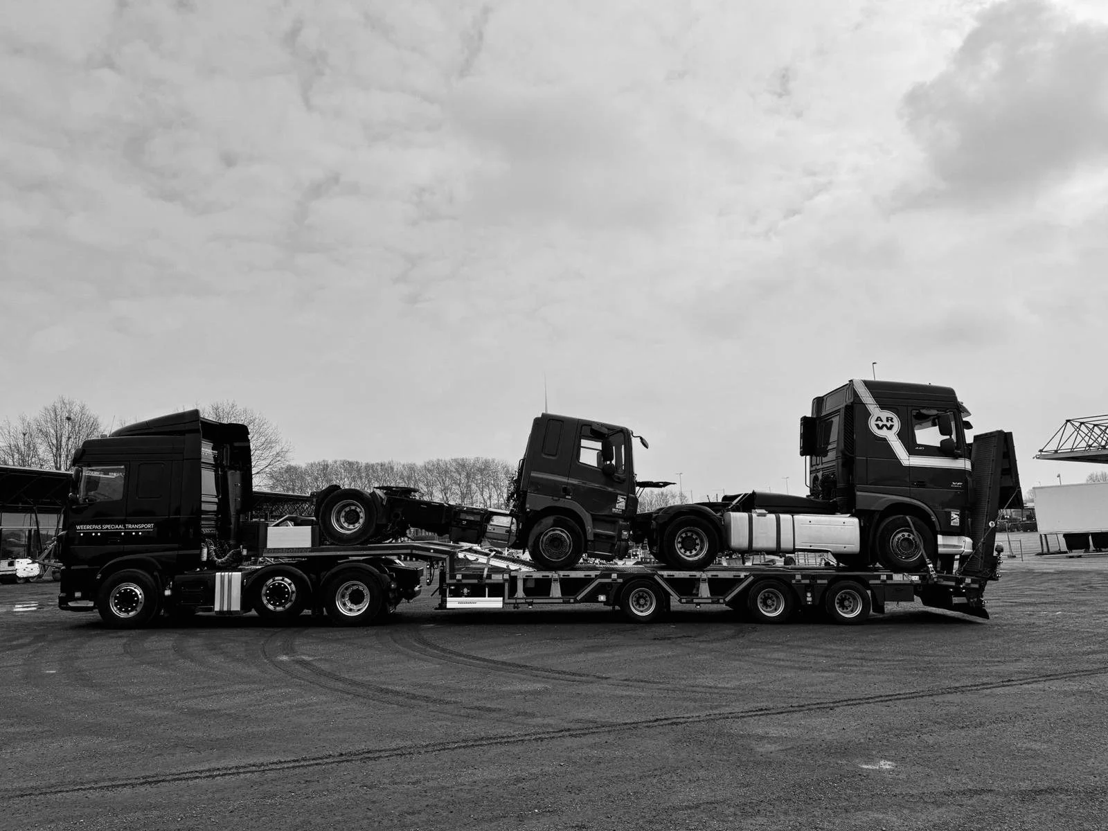 Zwarte transportwagen met twee trucks geladen met een truckcabine, op een parkeerterrein met bomen op de achtergrond, in zwart-wit