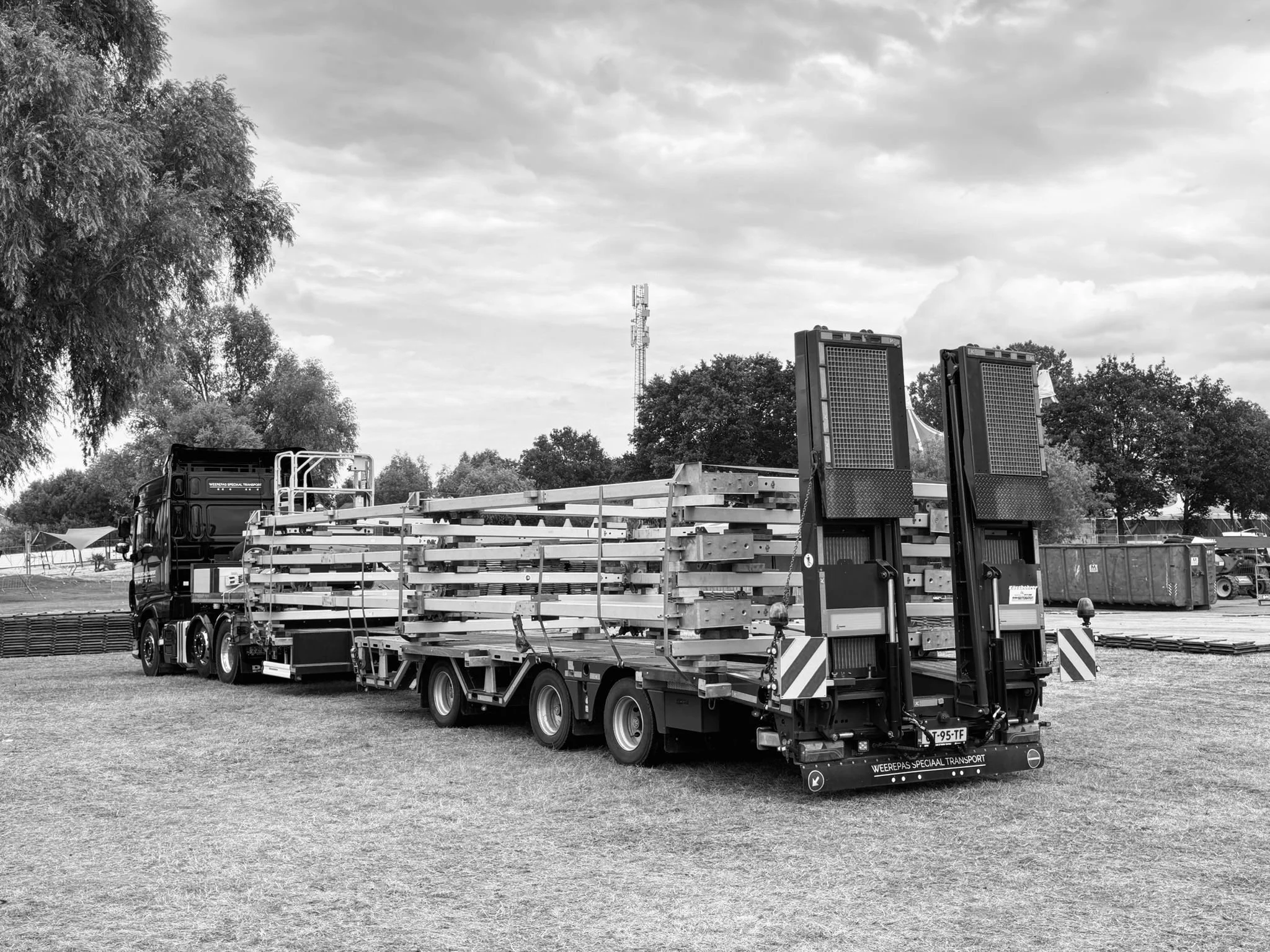 Transportvoertuig met grote machines en materialen op een grasveld, omringd door bomen, met een telecommunicatietoren op de achtergrond en een bewolkte hemel.