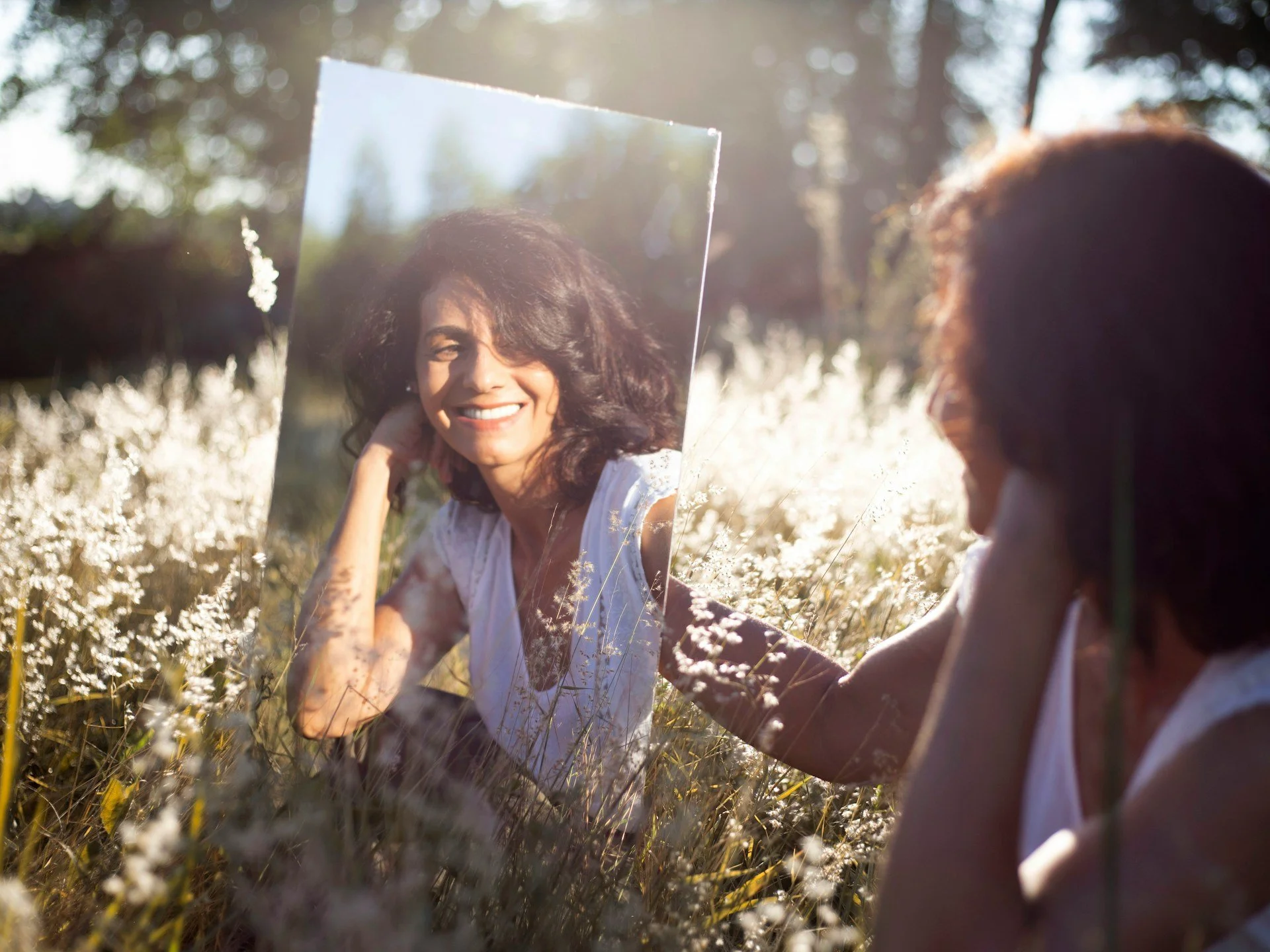 Femme de dos dans un champs de fleurs regardant son reflet dans un miroir, symbole de la reconstruction émotionnelle après un cancer