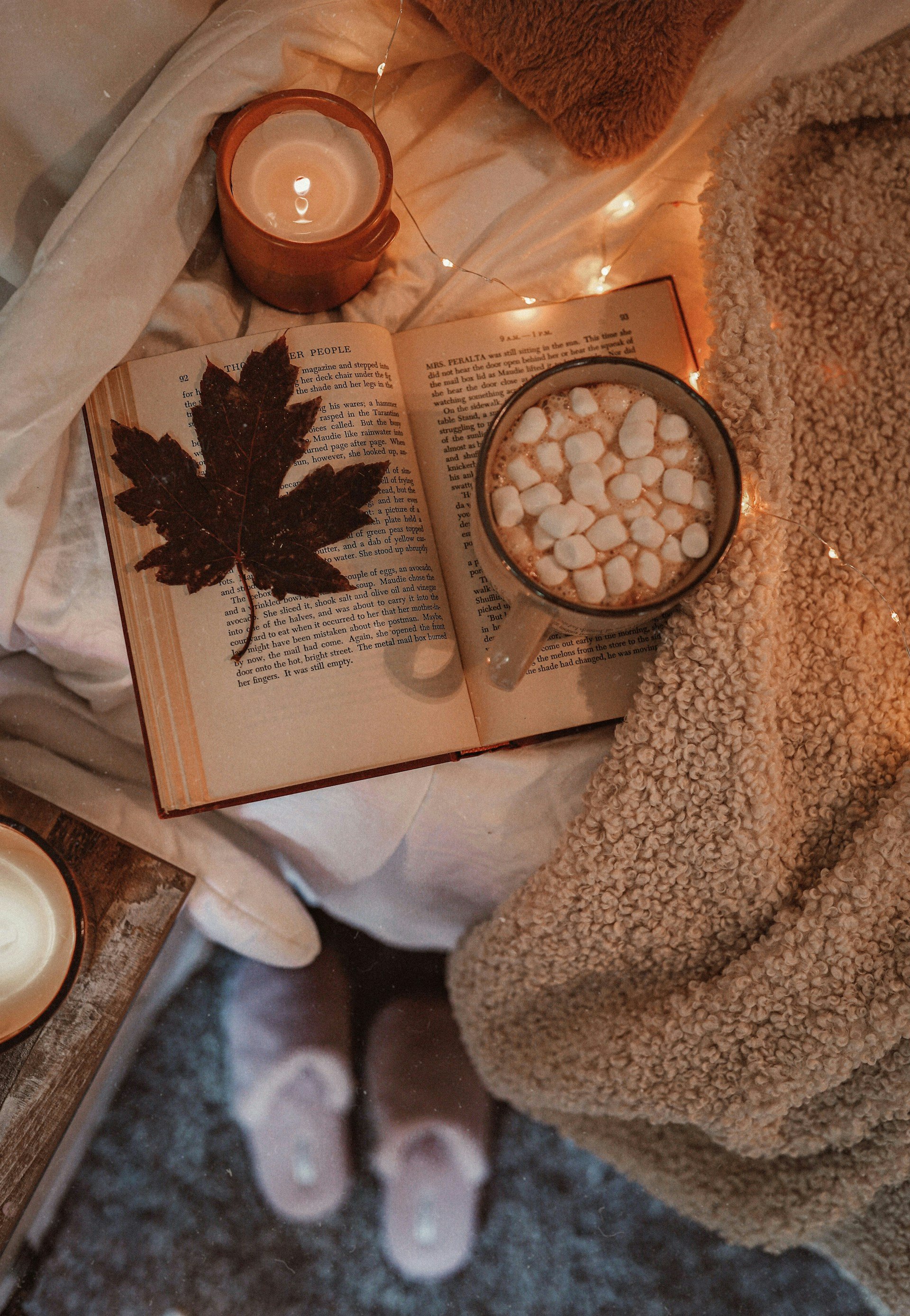livre ouvert avec une feuille d'arbre, tasse de marshmallows et bougie sur un lit, symbole de de réconfort et de bienveillance pendant les traitements contre le cancer