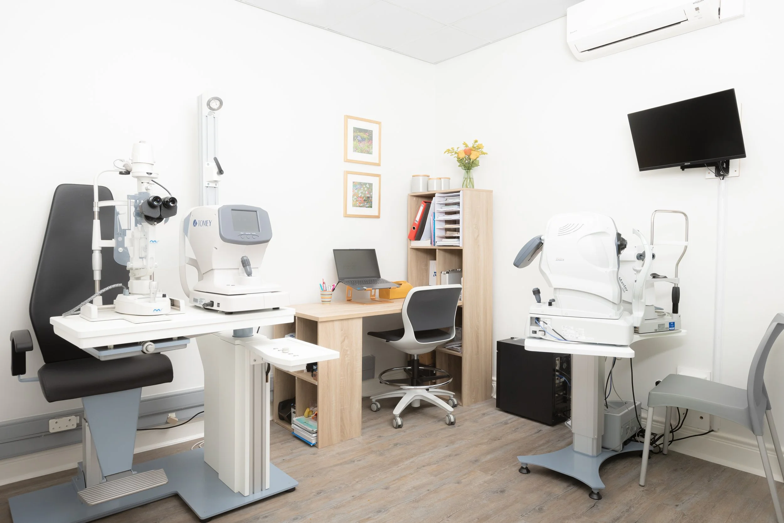 Medical examination room with ophthalmology equipment, computer, shelves, and a wall-mounted TV.