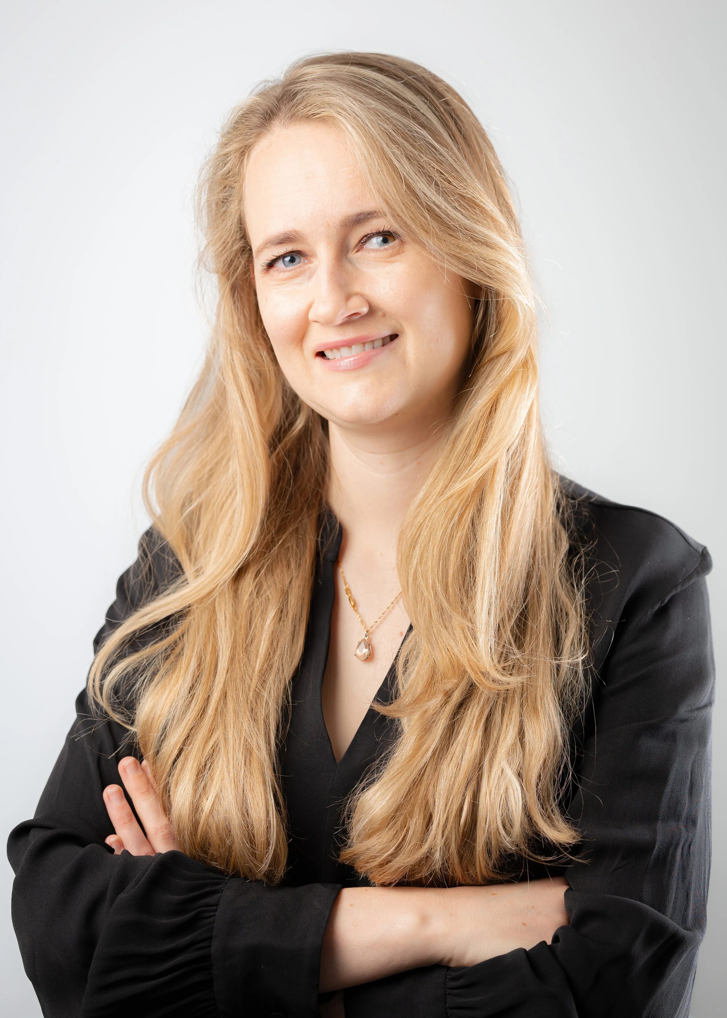 A woman with long blonde hair, blue eyes, wearing a black blouse and a pearl pendant necklace, standing with arms crossed against a plain white background.