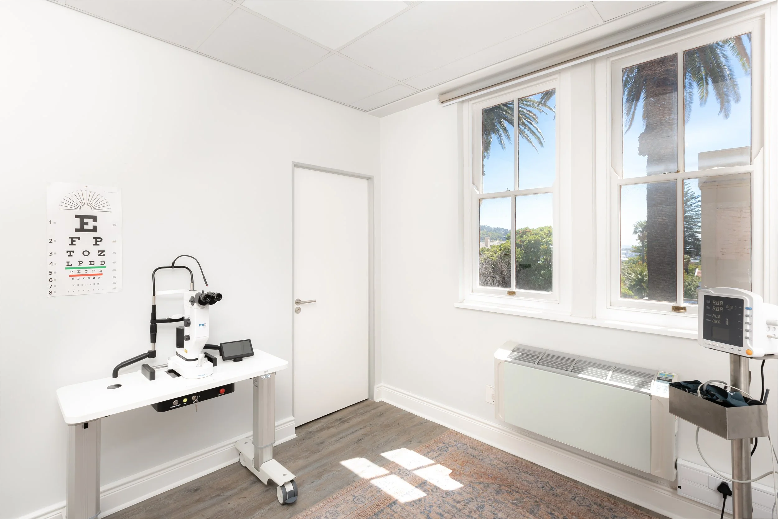 A medical examination room featuring a white cabinet with medical equipment including a microscope, ophthalmology eye chart on the wall, and a window with a view of palm trees outside, bright with natural sunlight.