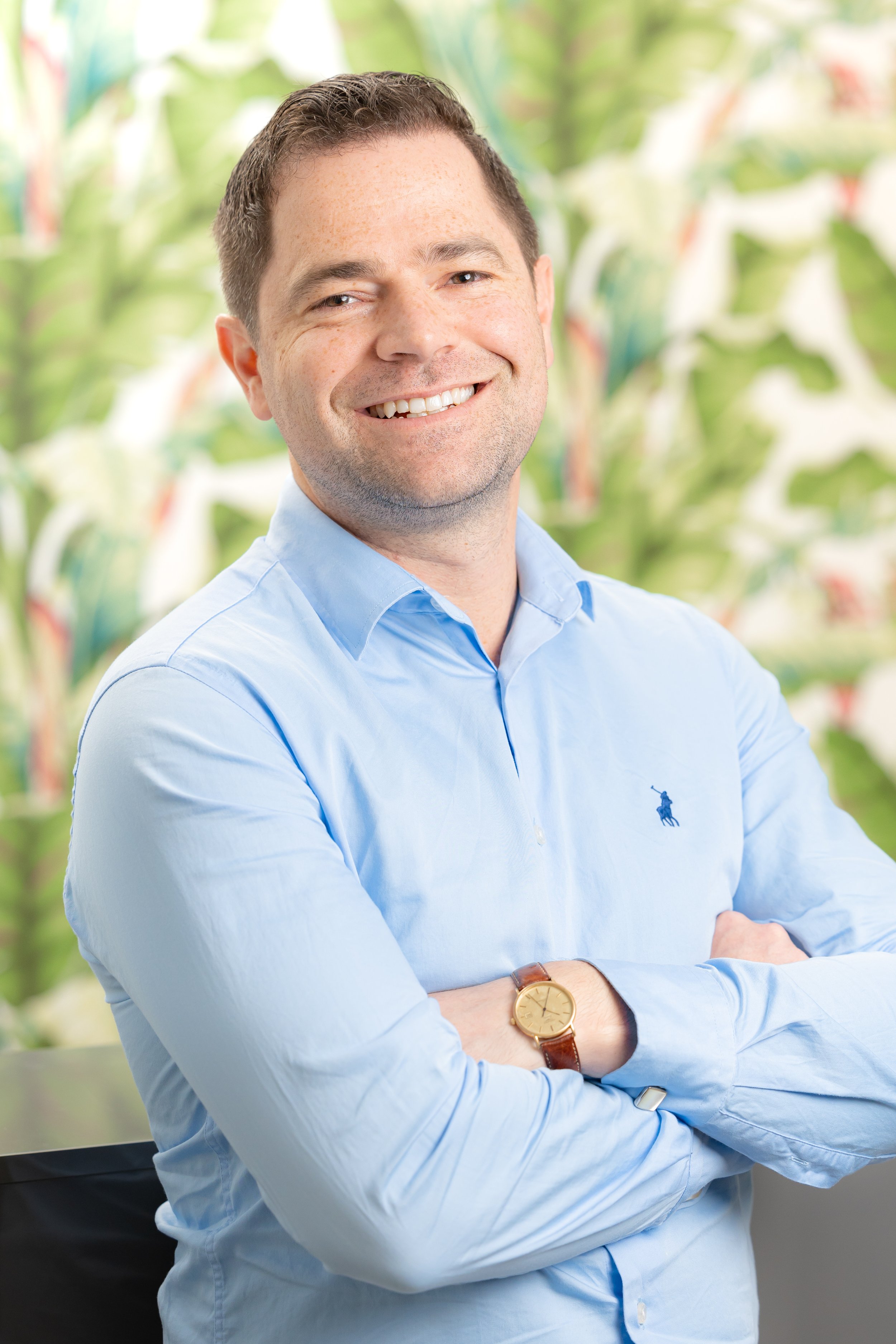 A smiling man with short brown hair and light skin, wearing a light blue button-up shirt and a gold wristwatch, standing with arms crossed in front of a green, leafy background.