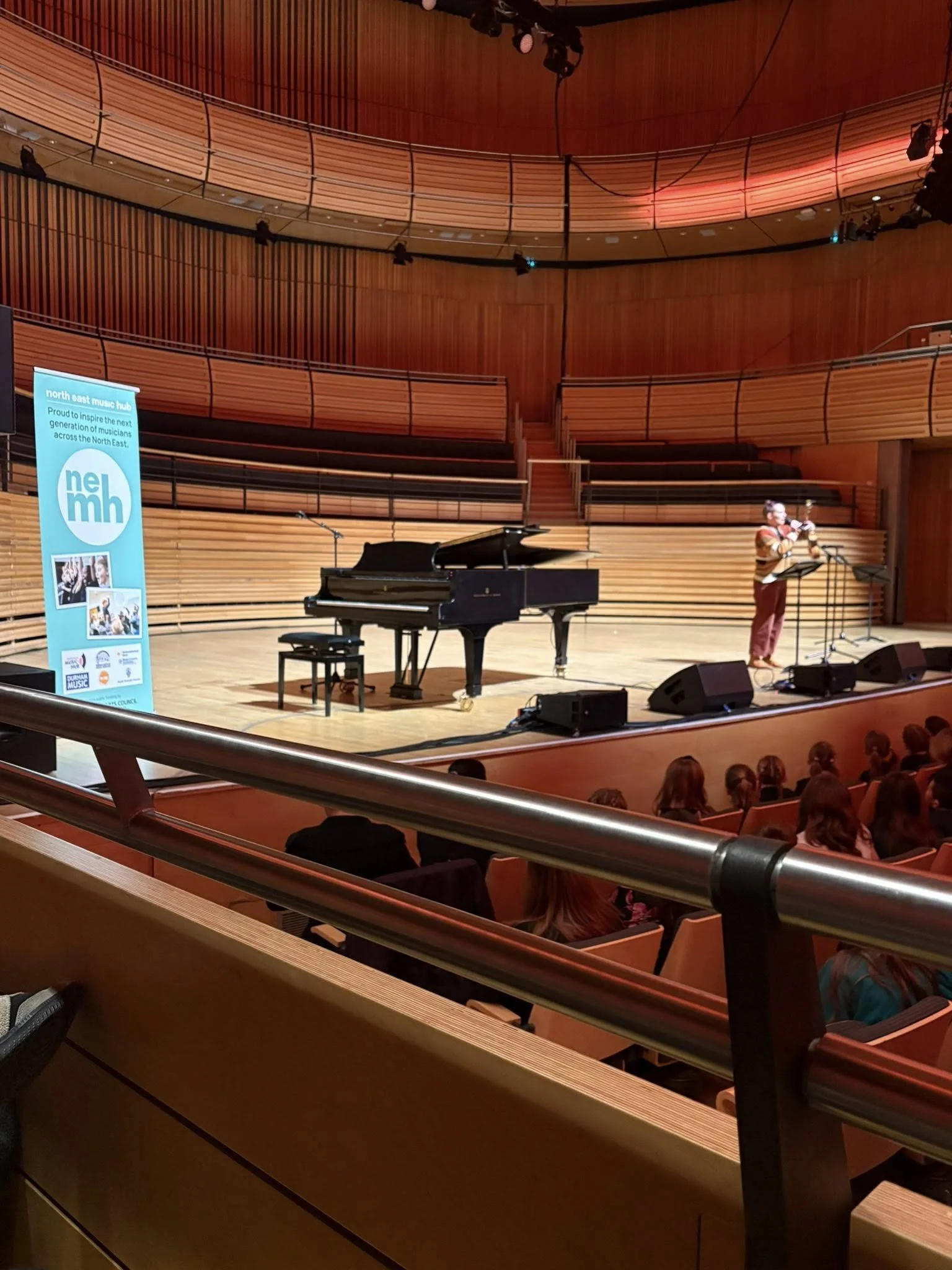 A lady standing on stage in front of a microphone with a ukulele in her hand, stood next to a grand piano and a north east music hub banner