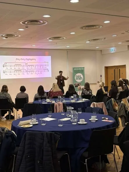 Facilitator standing at front of room with arms raised, with group of delegates sat in semi circle facing them.