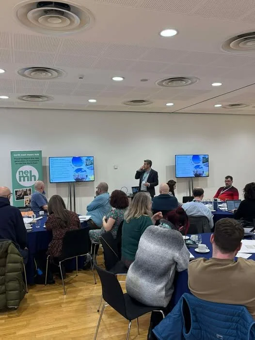 Man standing at the front of the room presenting to men and women sat at round tables.